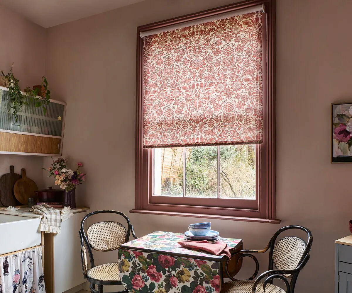 Kitchen dining nook with a window framed in wood, dressed with a patterned Roller blind that filters soft daylight above a small table.