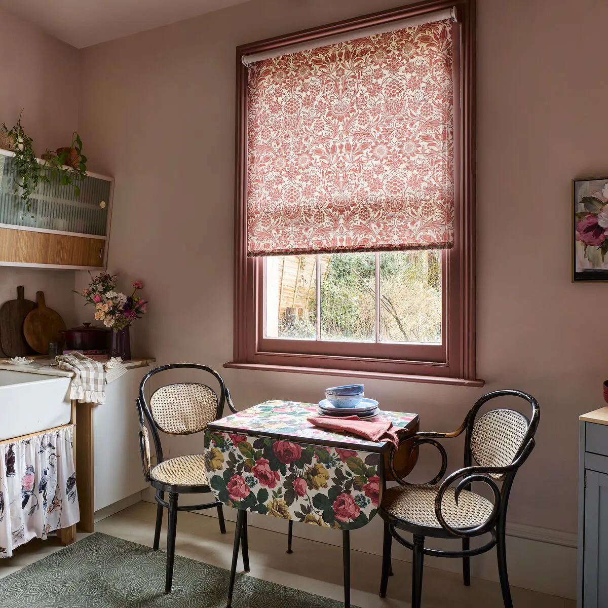 Kitchen dining nook with a window framed in wood, dressed with a patterned Roller blind that filters soft daylight above a small table.