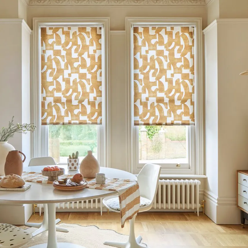 Bright dining room with two tall sash windows dressed in patterned Roller blinds, letting in soft daylight above a white table.