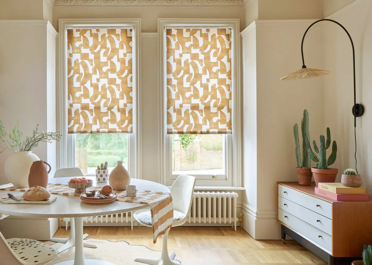 Bright dining room with two tall sash windows dressed in patterned Roller blinds, letting in soft daylight above a white table.