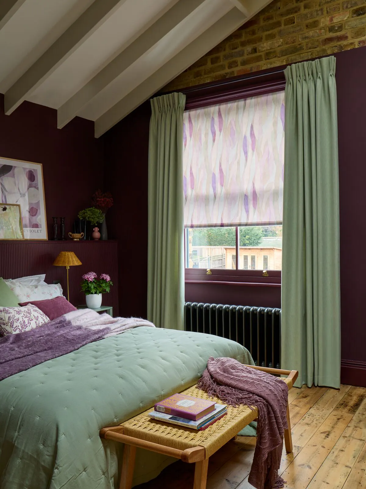 Bedroom with exposed beams and burgundy walls, window with patterned Roller blind in amethyst tones and sage curtains over radiator.