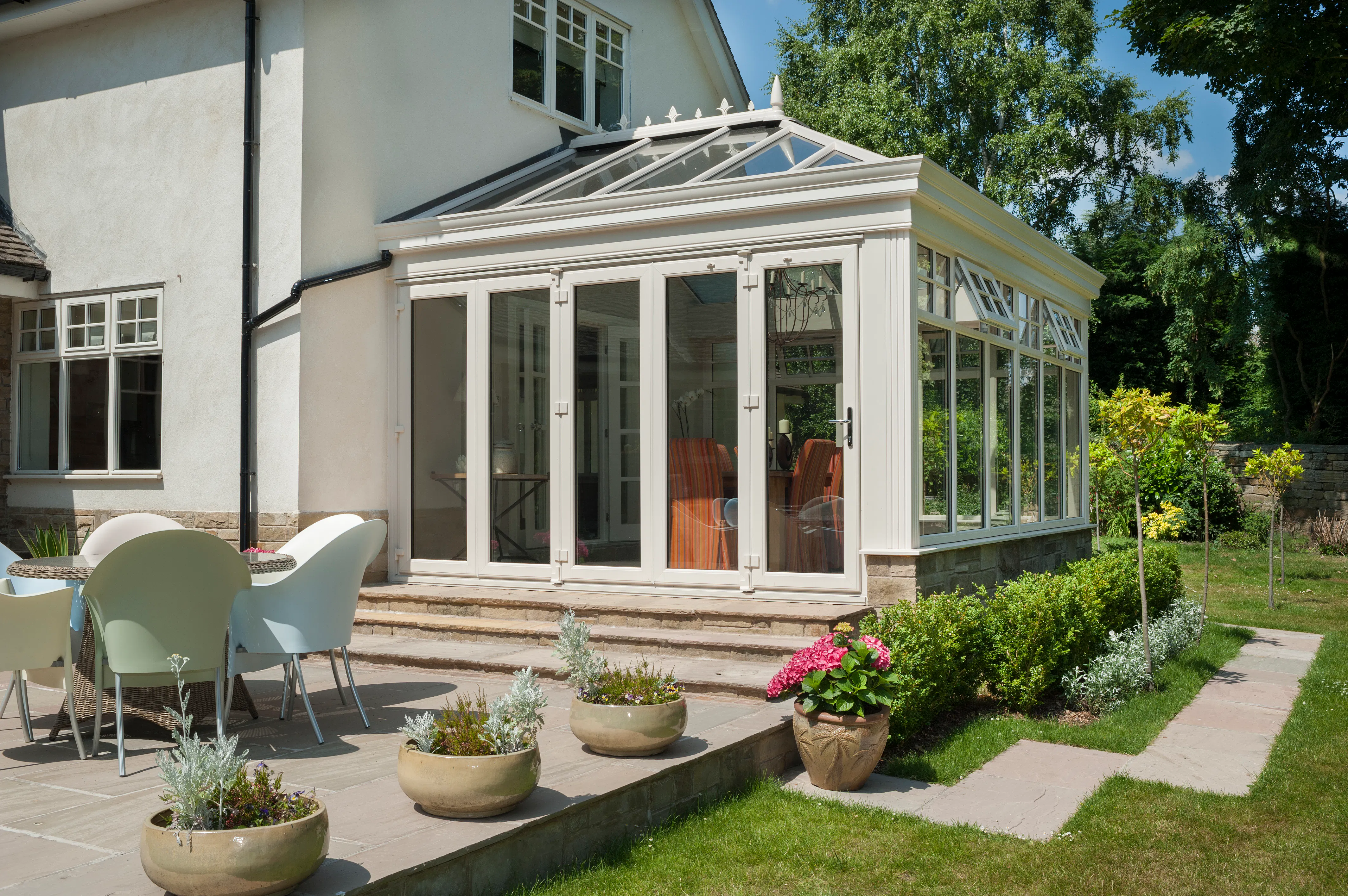 White conservatory with glazed roof and tall glass doors, set on a patio with potted plants and lawn beyond.