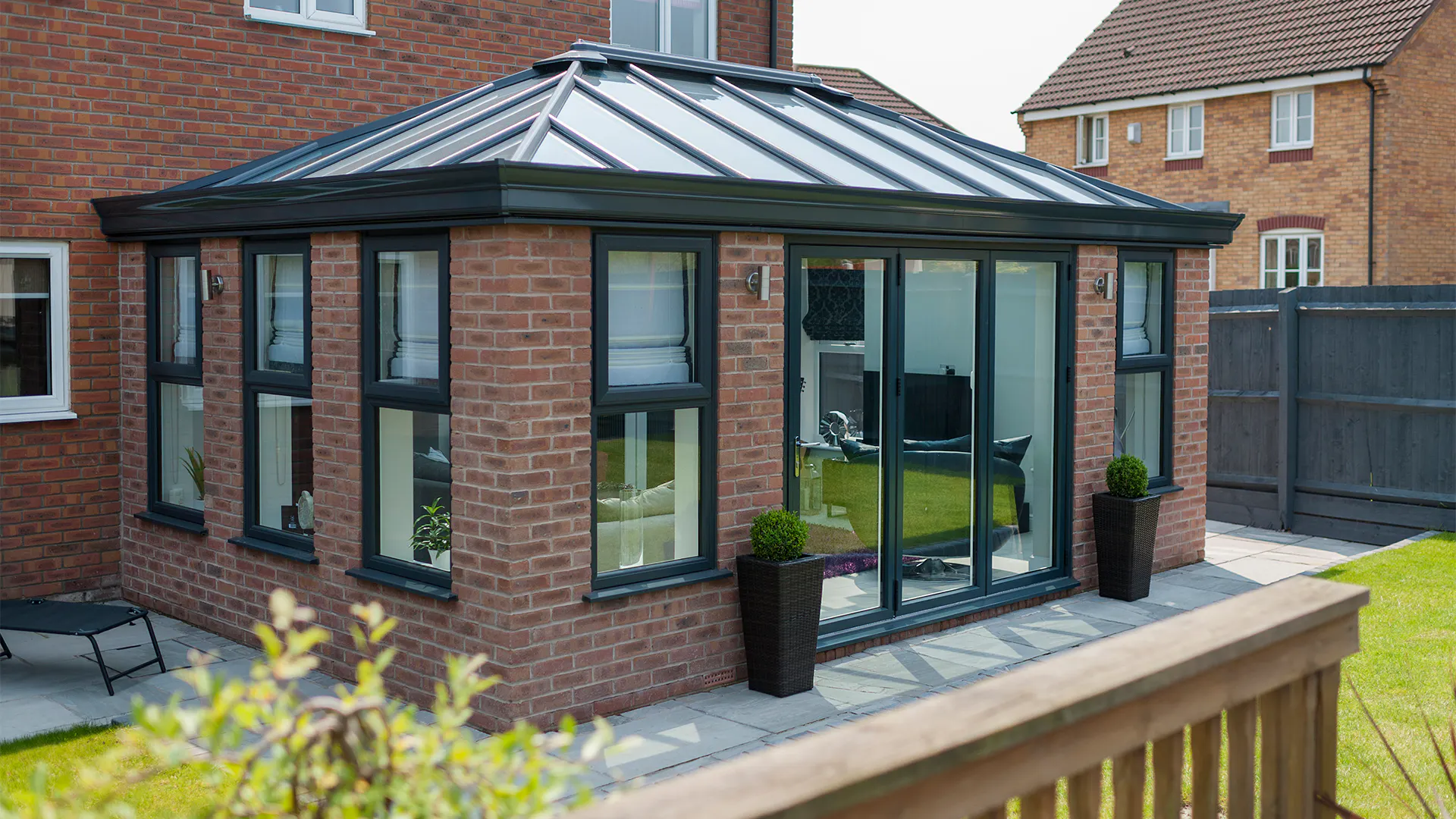 Brick-built orangery with dark-framed glazing and a hipped roof, opening onto a paved terrace beside a fenced lawn.