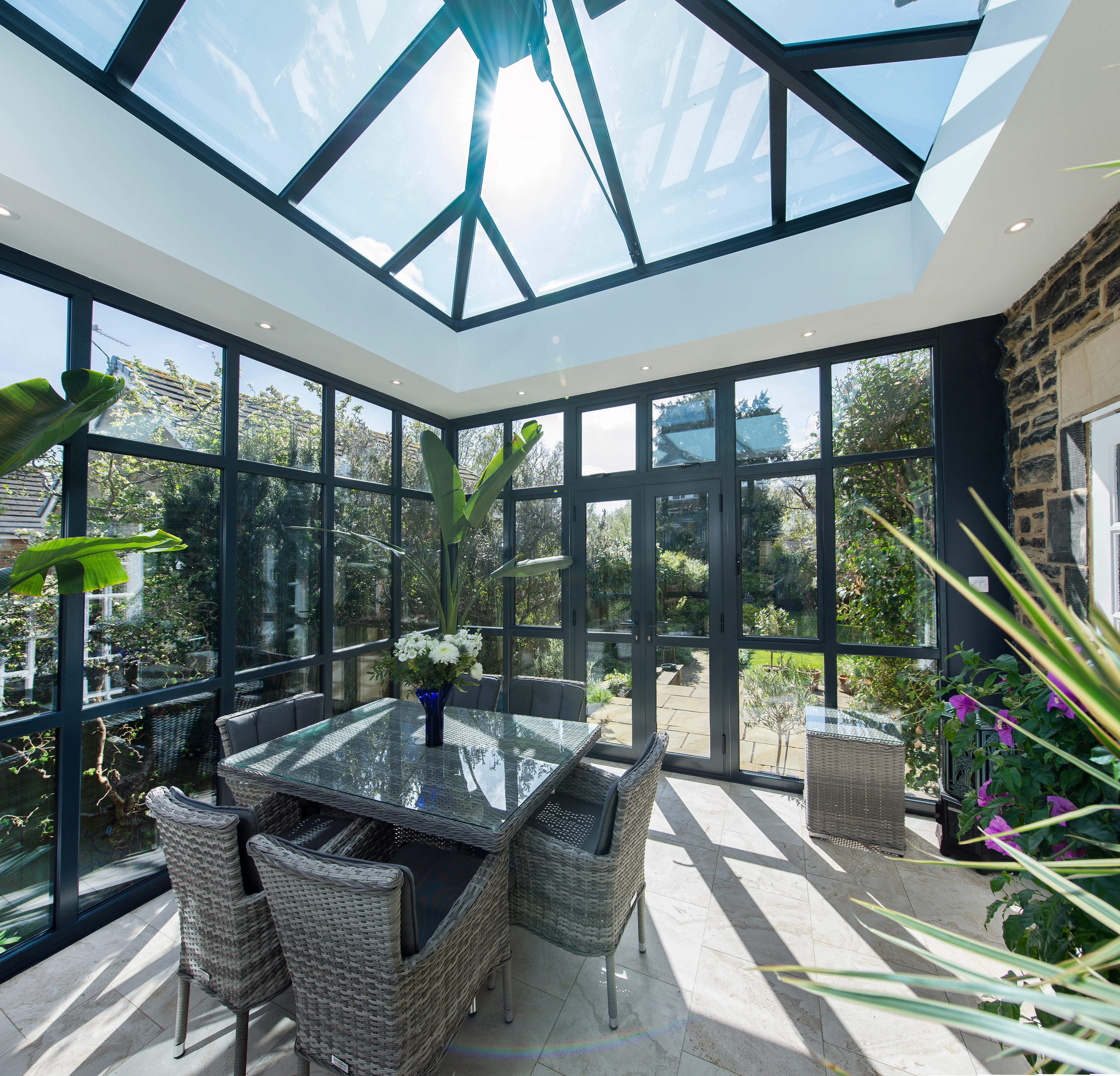 Light-filled garden room with black-framed roof lantern and floor-to-ceiling windows, creating an airy dining space overlooking greenery.