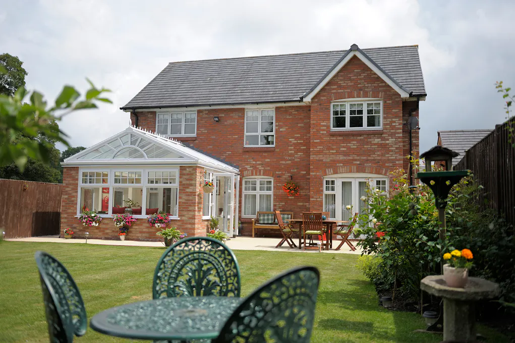 Brick home with white-framed conservatory featuring a glass roof, surrounded by hanging baskets and potted plants in a neat garden.