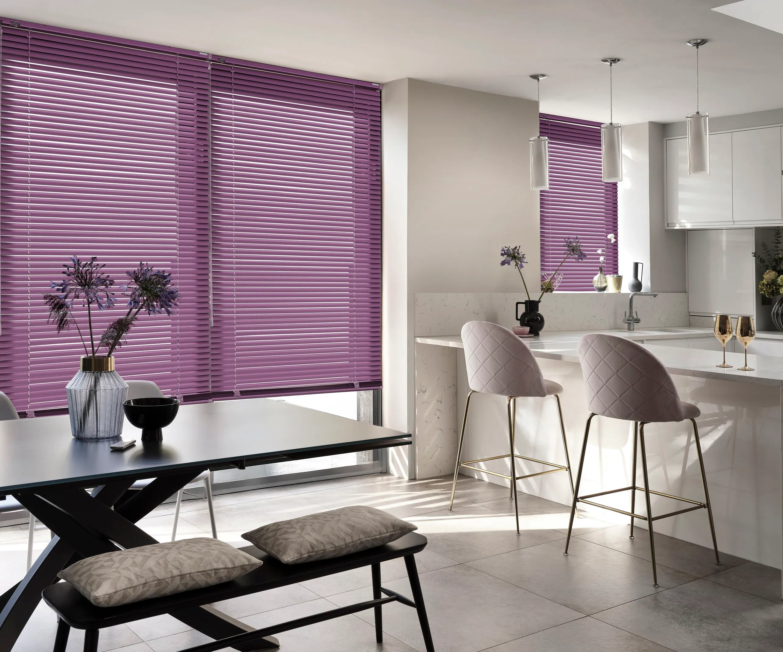 A modern open-plan kitchen and dining room featuring violet-coloured metal Venetian blinds on large windows, paired with white cabinetry and grey floor tiles.