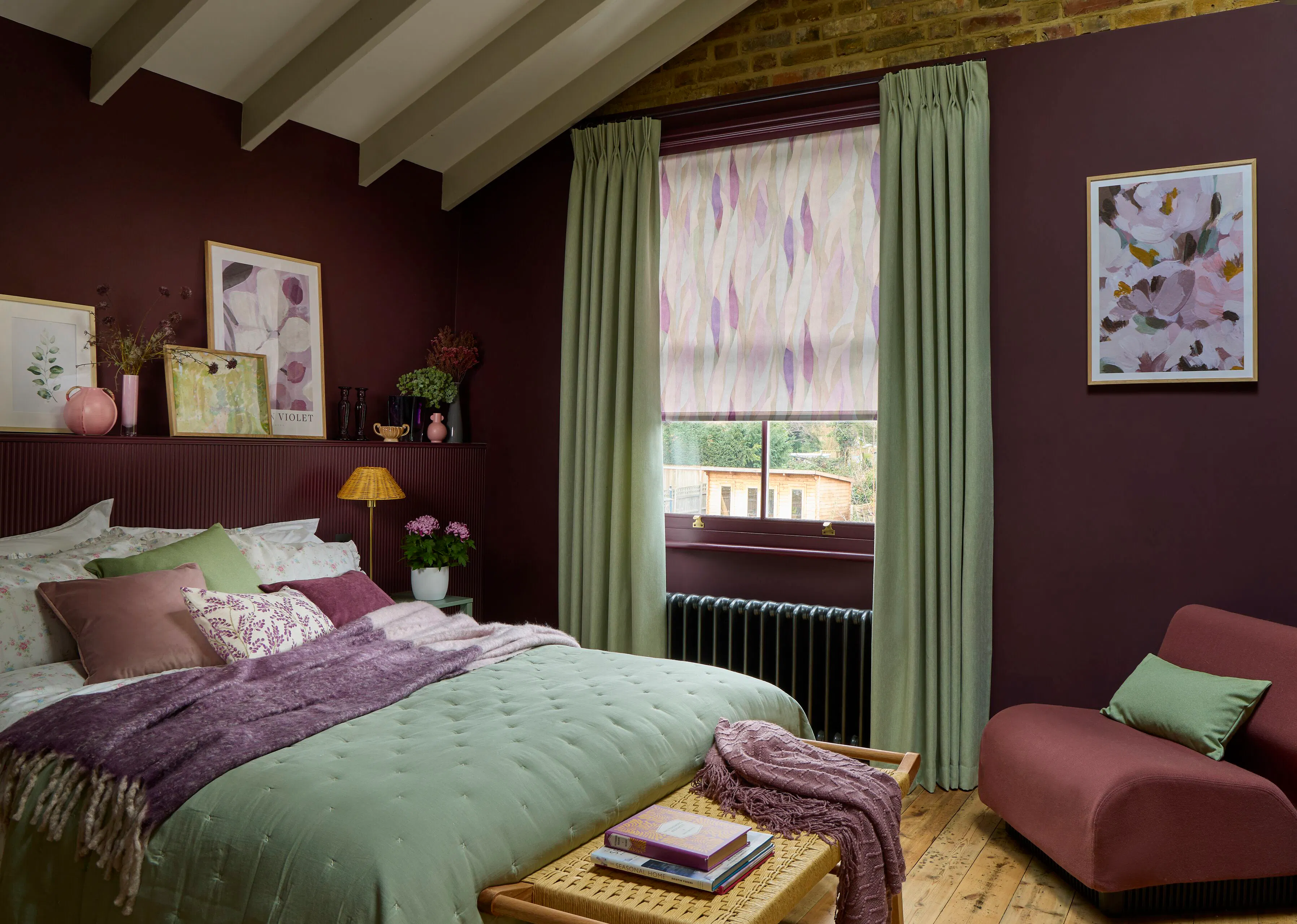 A cosy bedroom with deep-plum walls, sage-green curtains and a purple and pink Roller blind, styled with coordinating bedding in soft greens, mauves and pinks.