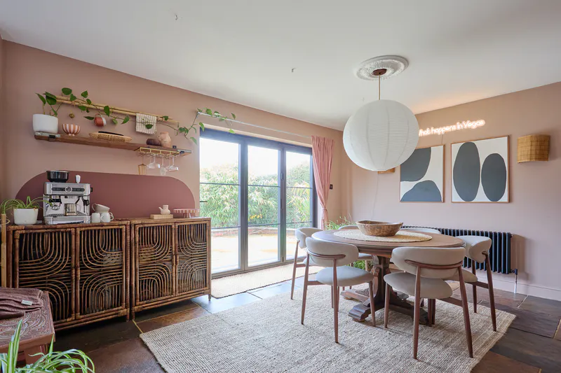 Dining space featuring wide glass patio doors framed by soft pink curtains, opening the room to garden light and greenery.