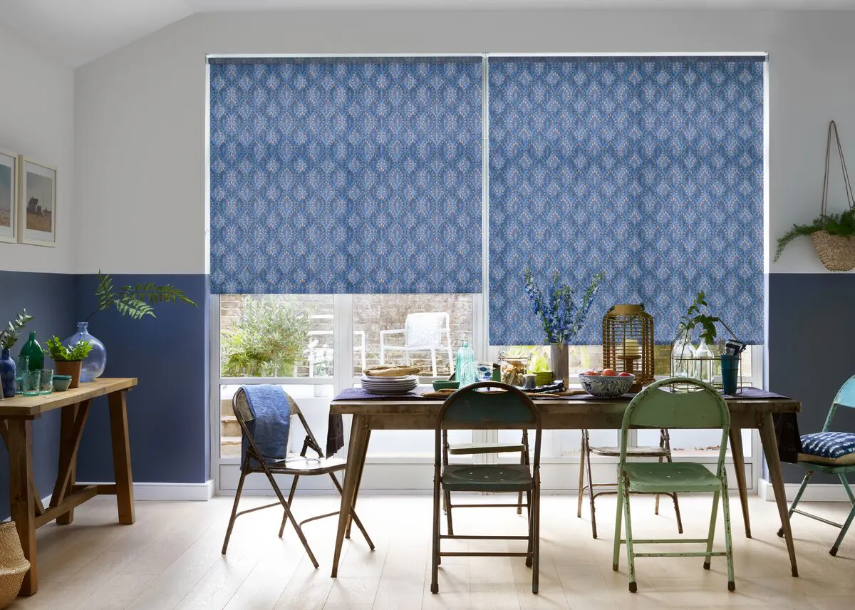 Dining room window with patterned blue Roller blinds partly lowered over glazed doors, letting in soft daylight above a wooden table setting.