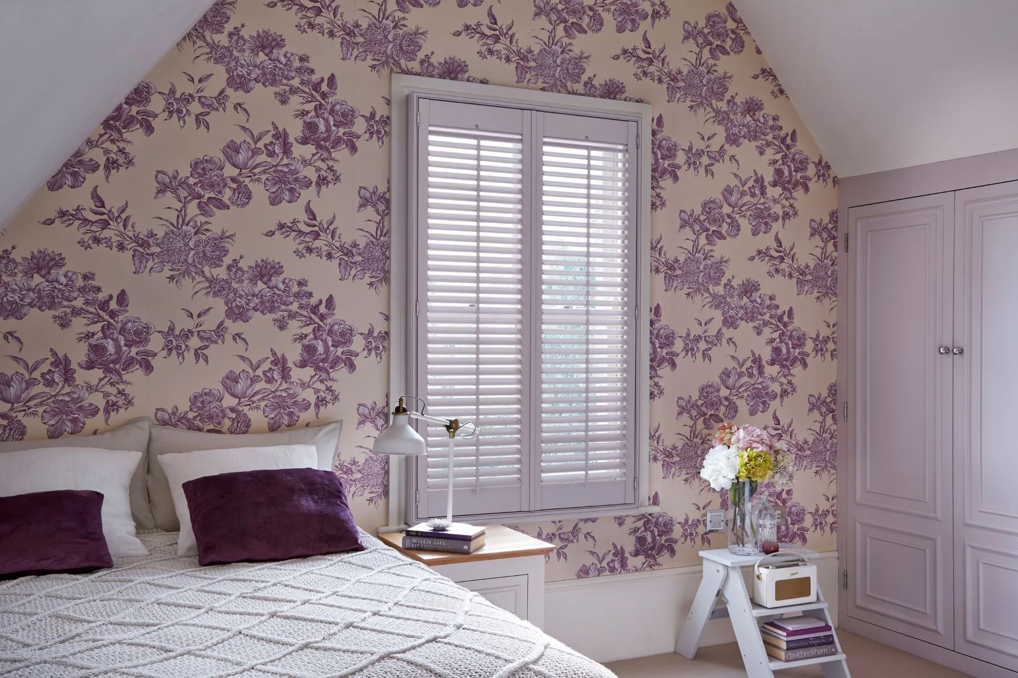 A romantic loft bedroom with soft purple and plaster-pink floral wallpaper, lilac-painted shutters and white bedding accented with deep plum cushions.