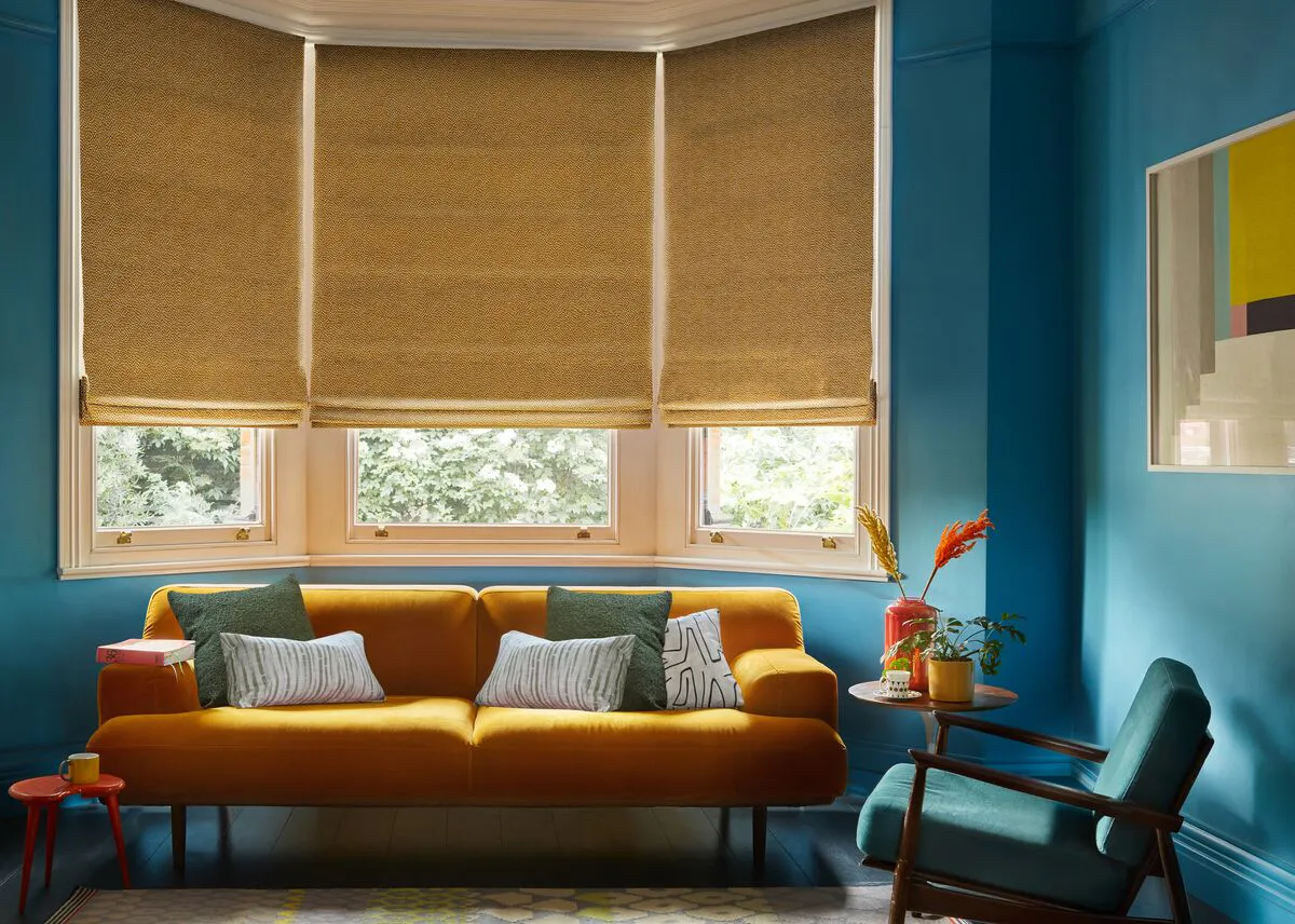 A vibrant teal living room with three golden, textured Roman blinds in a bay window and a rich mustard velvet sofa.