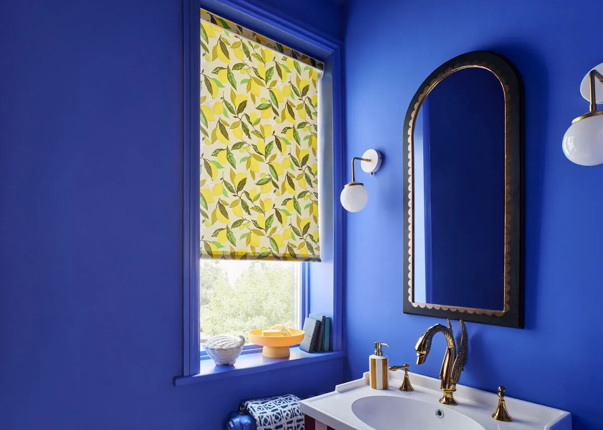 A bold, Mediterranean-inspired cobalt blue bathroom with a vibrant lemon print Roller blind, arched mirror and brass fixtures.