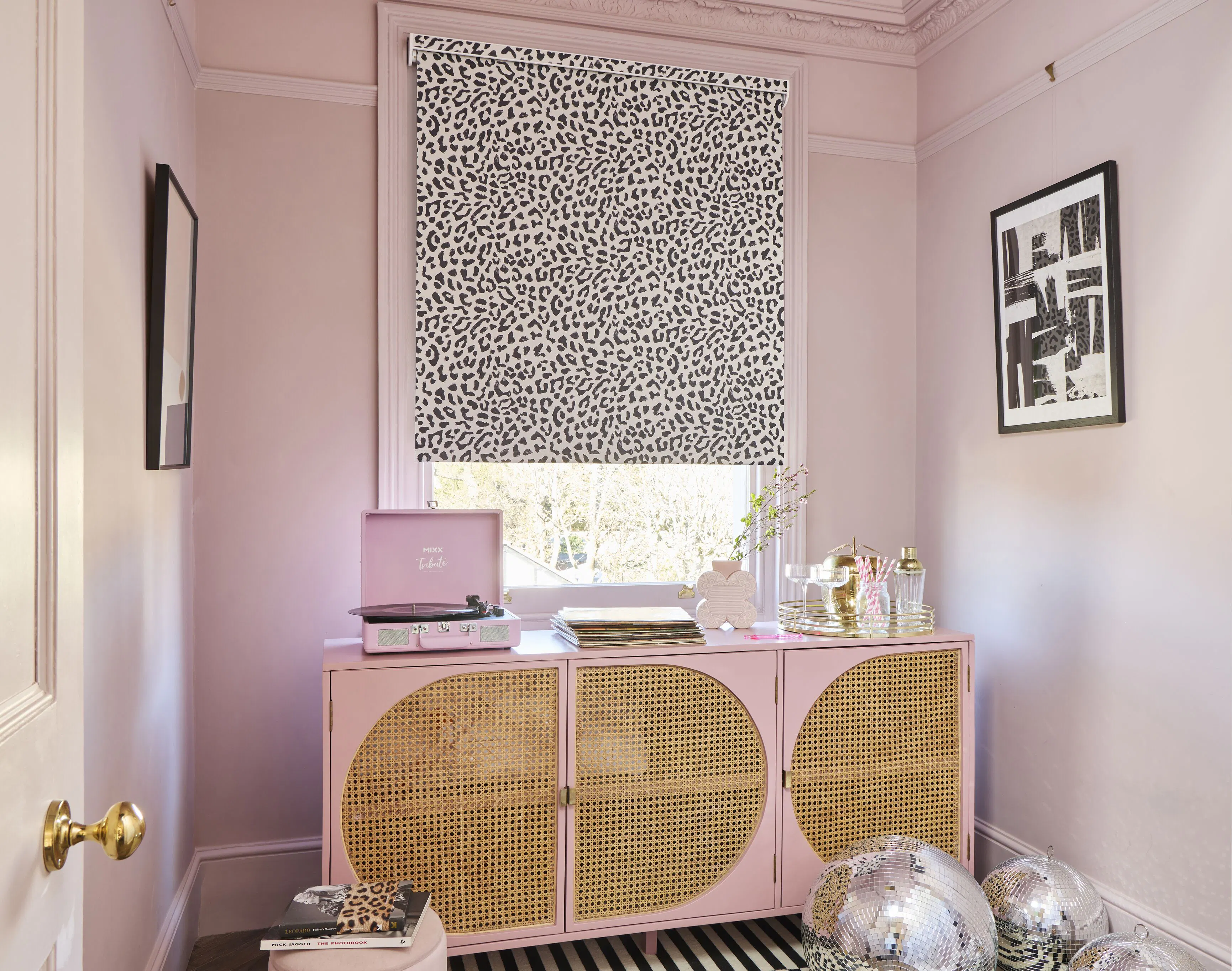 A colour-drenched pink dressing room featuring a monochrome leopard print Roller blind above a rattan-fronted sideboard and cluster of disco balls.