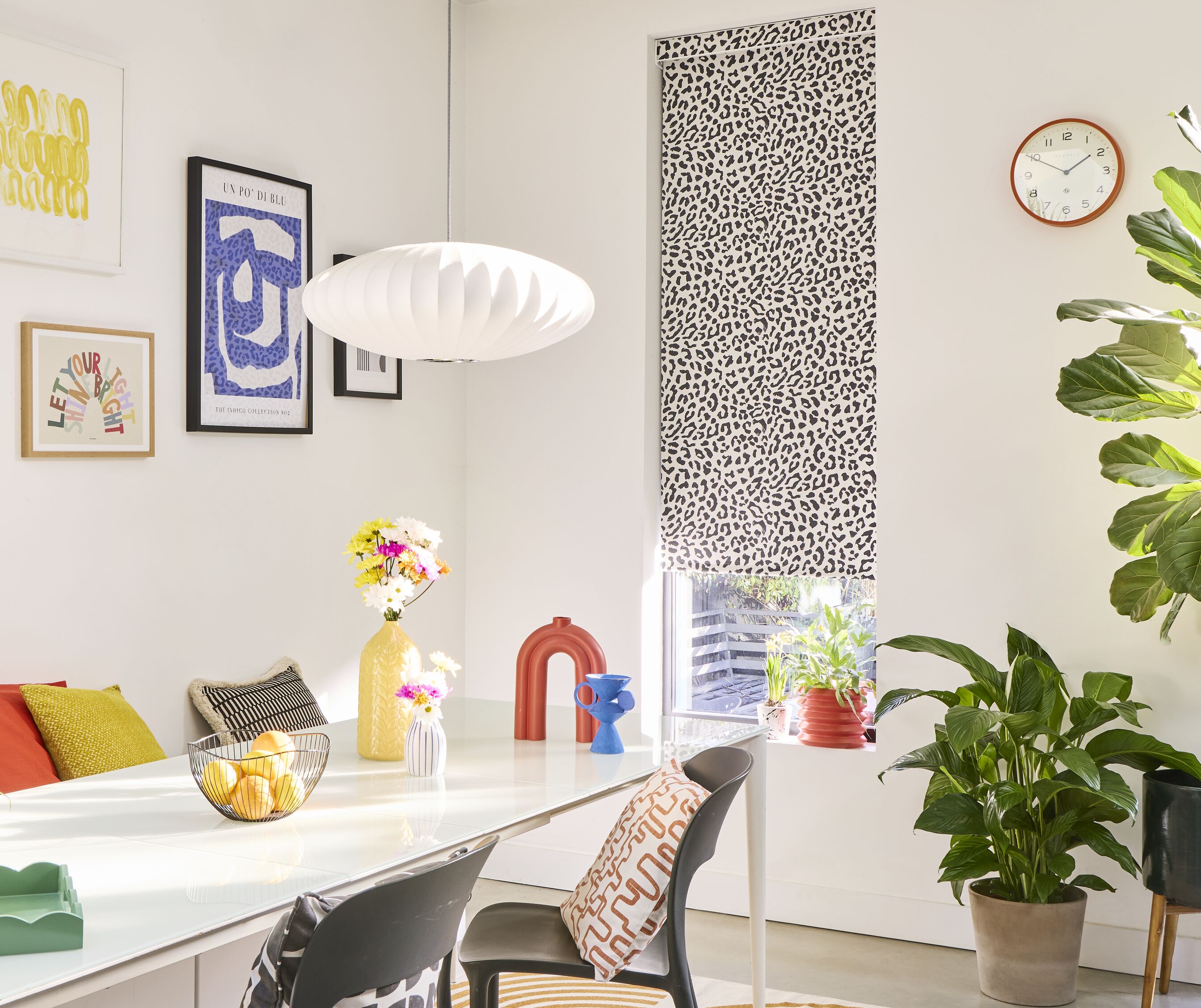 A bright, white-walled dining area with a monochrome leopard print Roller blind, styled with colourful wall art and accessories and leafy houseplants.
