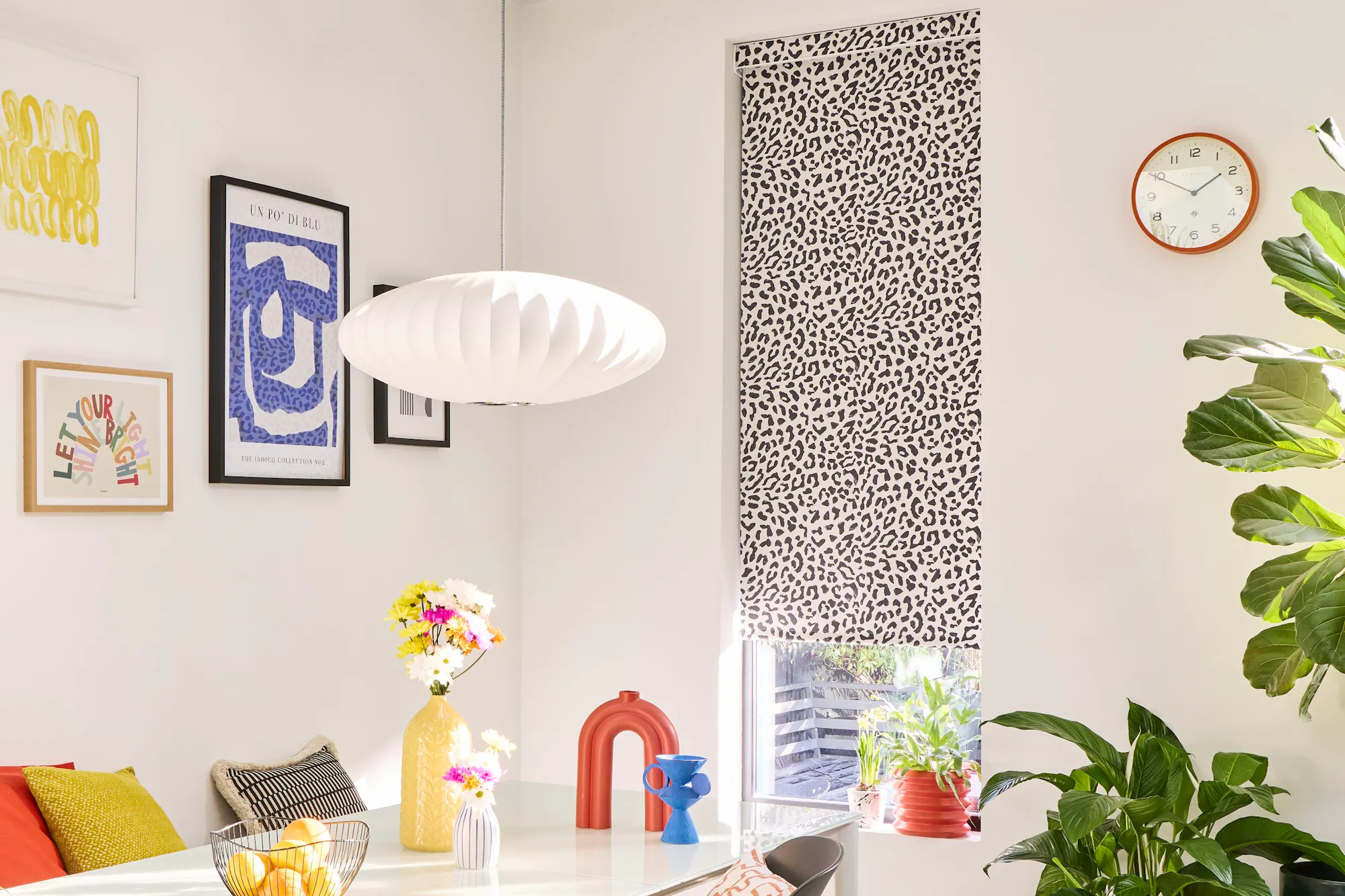A bright, white-walled dining area with a monochrome leopard print Roller blind, styled with colourful wall art and accessories and leafy houseplants.