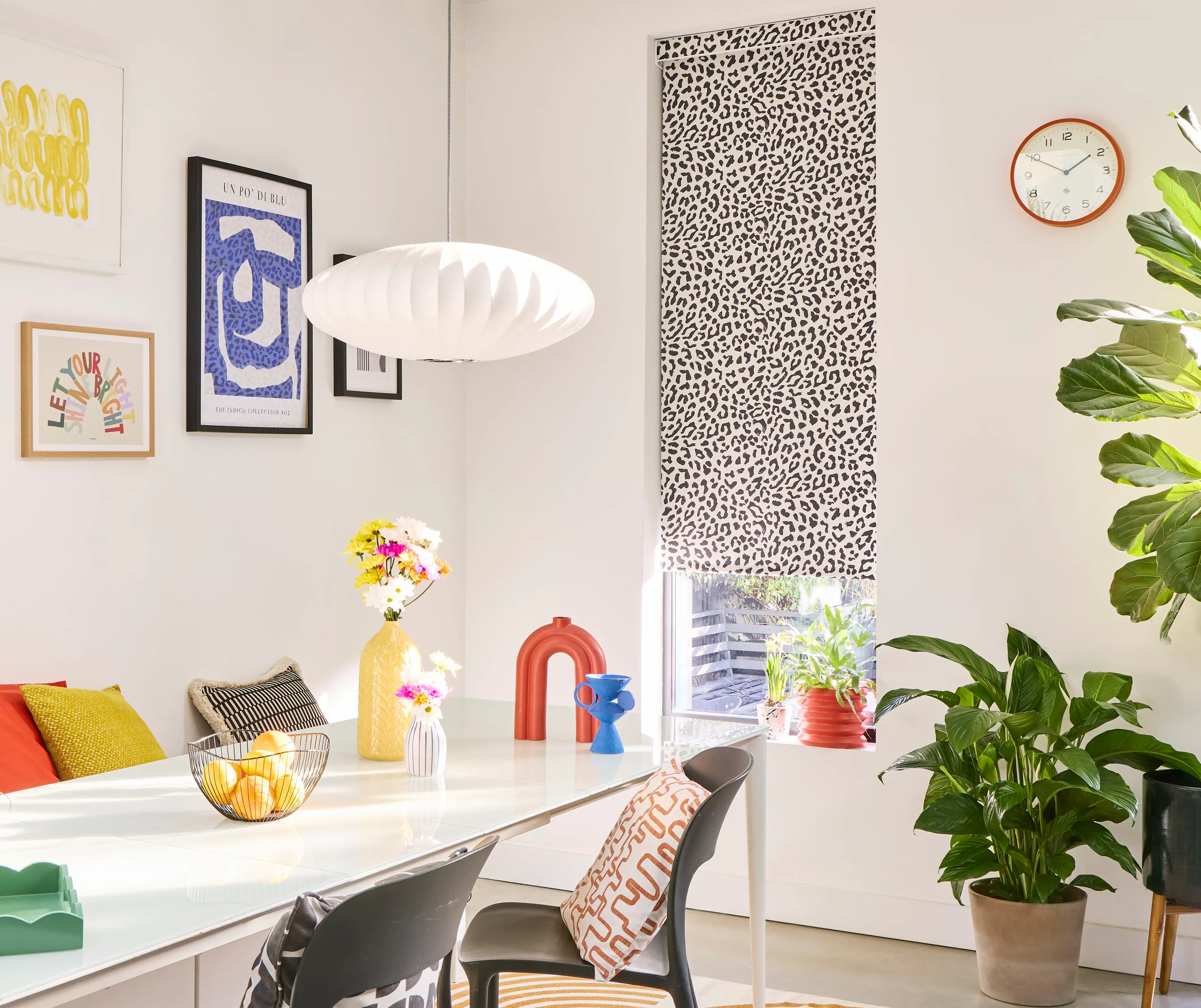 A bright, white-walled dining area with a monochrome leopard print Roller blind, styled with colourful wall art and accessories and leafy houseplants.