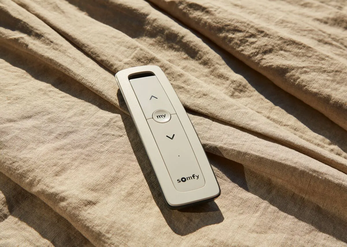 Somfy remote control resting on textured beige fabric, with simple up, down and “my” buttons centred in a slim white design.