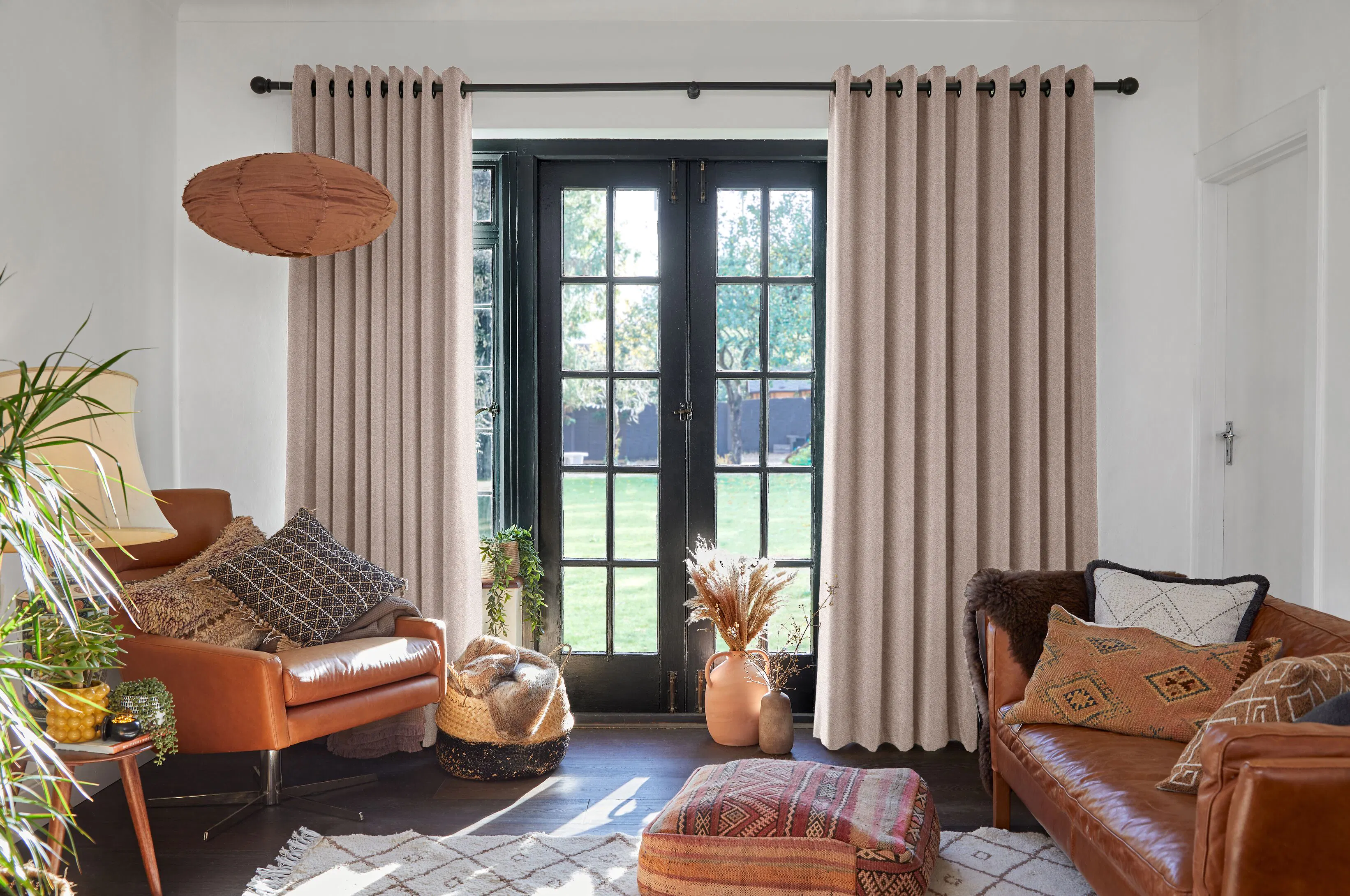 A light, boho-style garden room with tan leather sofas, heaped cushions and dried grasses. Floor-length natural curtains hang either side of black-framed French doors and a sunny garden is visible outside.