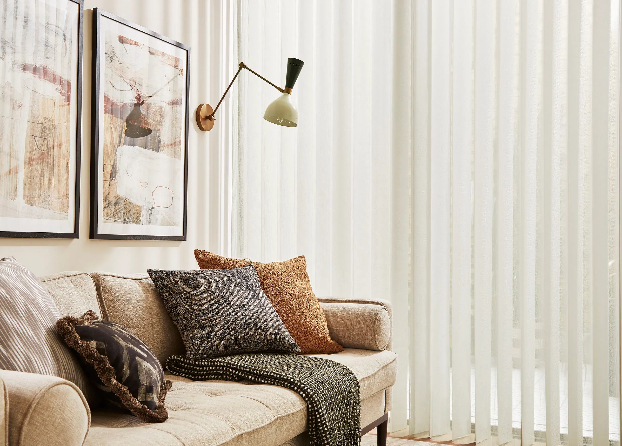 A neutral-toned corner of a room with sheer white Diffused blinds, a cream sofa with textured cushions and a mid-century style wall lamp.