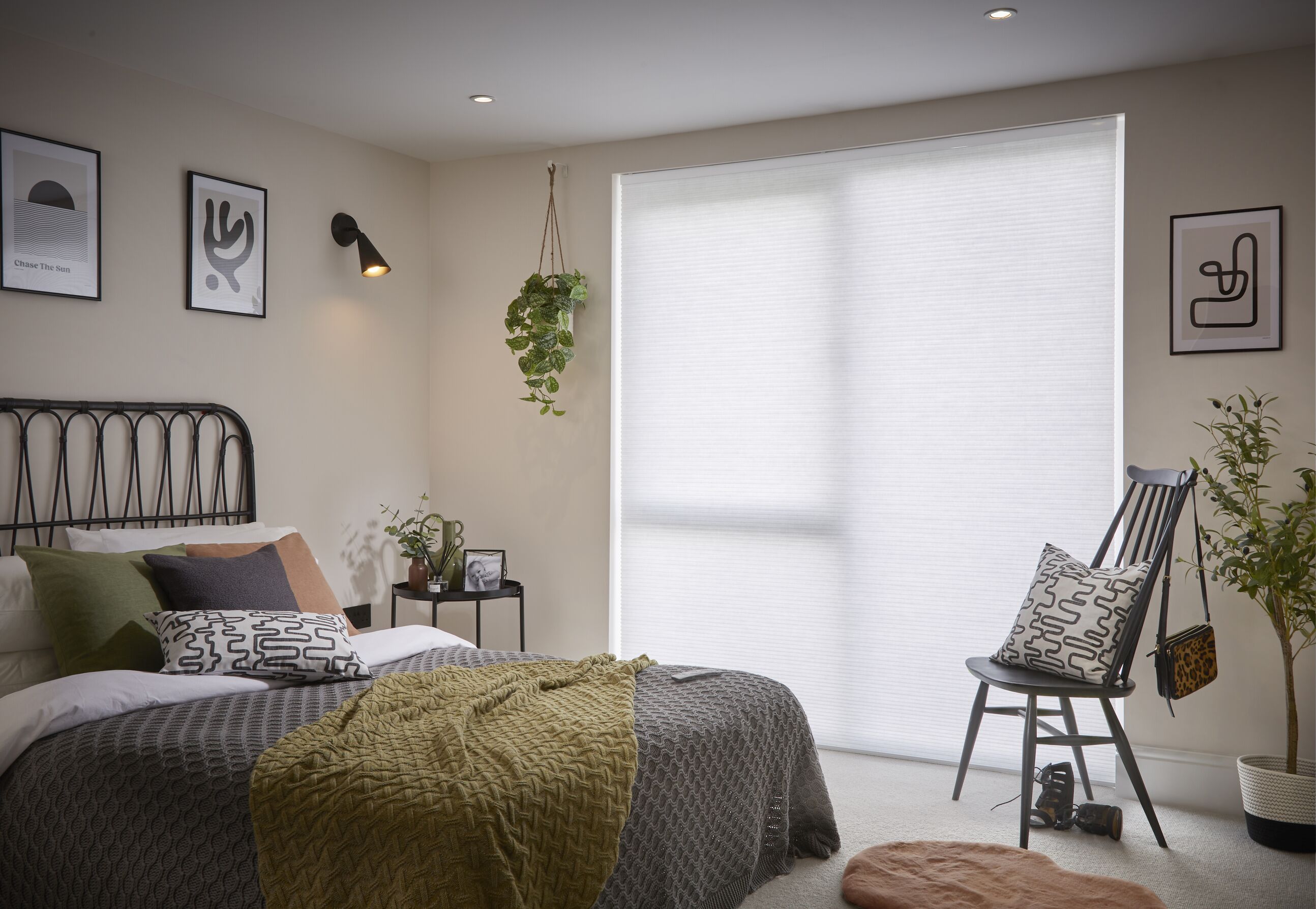 A cosy bedroom with pale plaster-coloured walls, a large white blind on a floor-to-ceiling window, black iron bed frame and layered olive green and charcoal bedding.