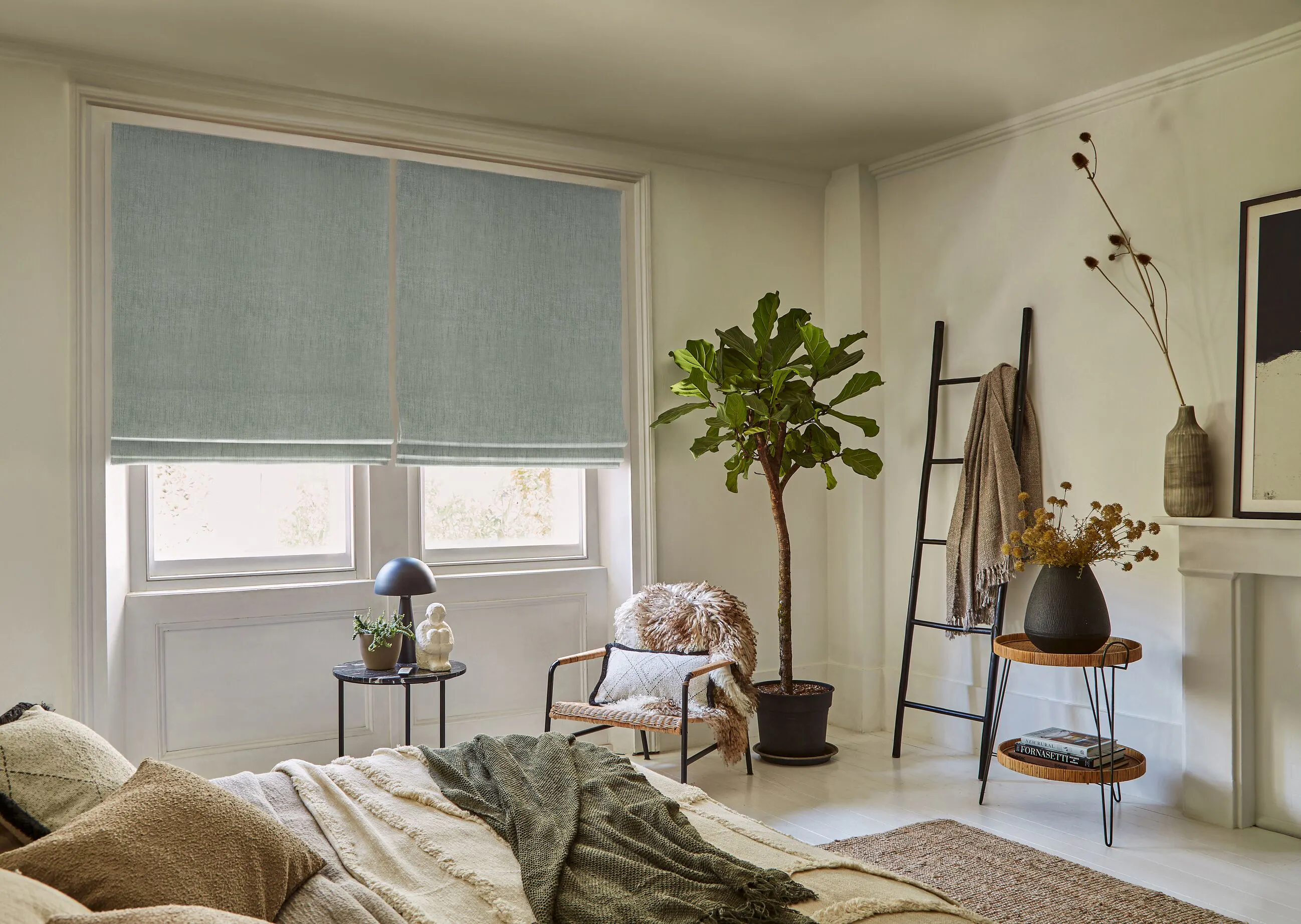 A calm, airy bedroom with two sage green blinds on a large sash window. With layered bedding, a fiddle leaf fig tree and a decorative wooden ladder.
