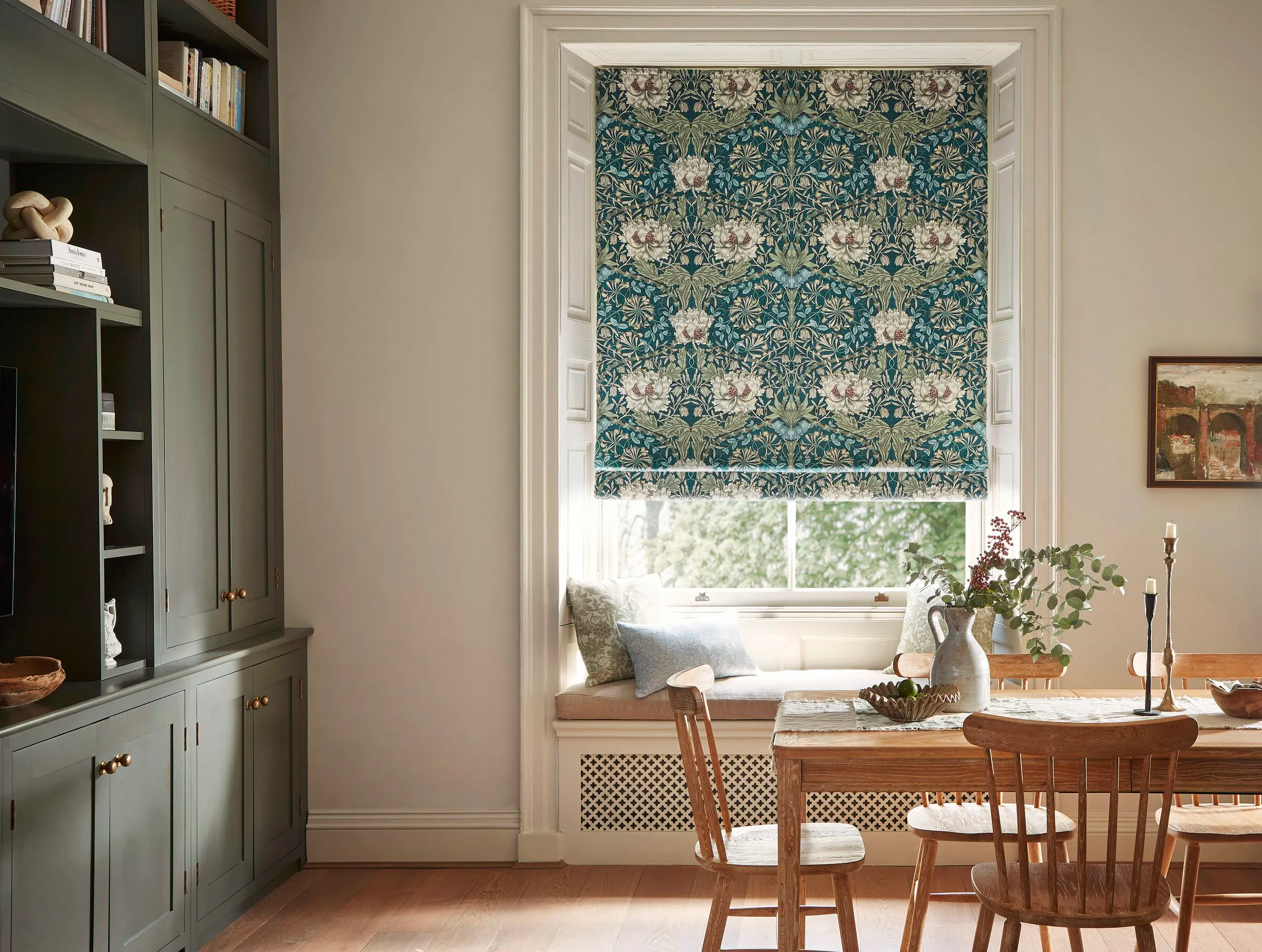 A light, airy dining with a farmhouse-style wooden table and chairs, tall grey-green painted cupboards and a William Morris Roman blind in a bay window seat.