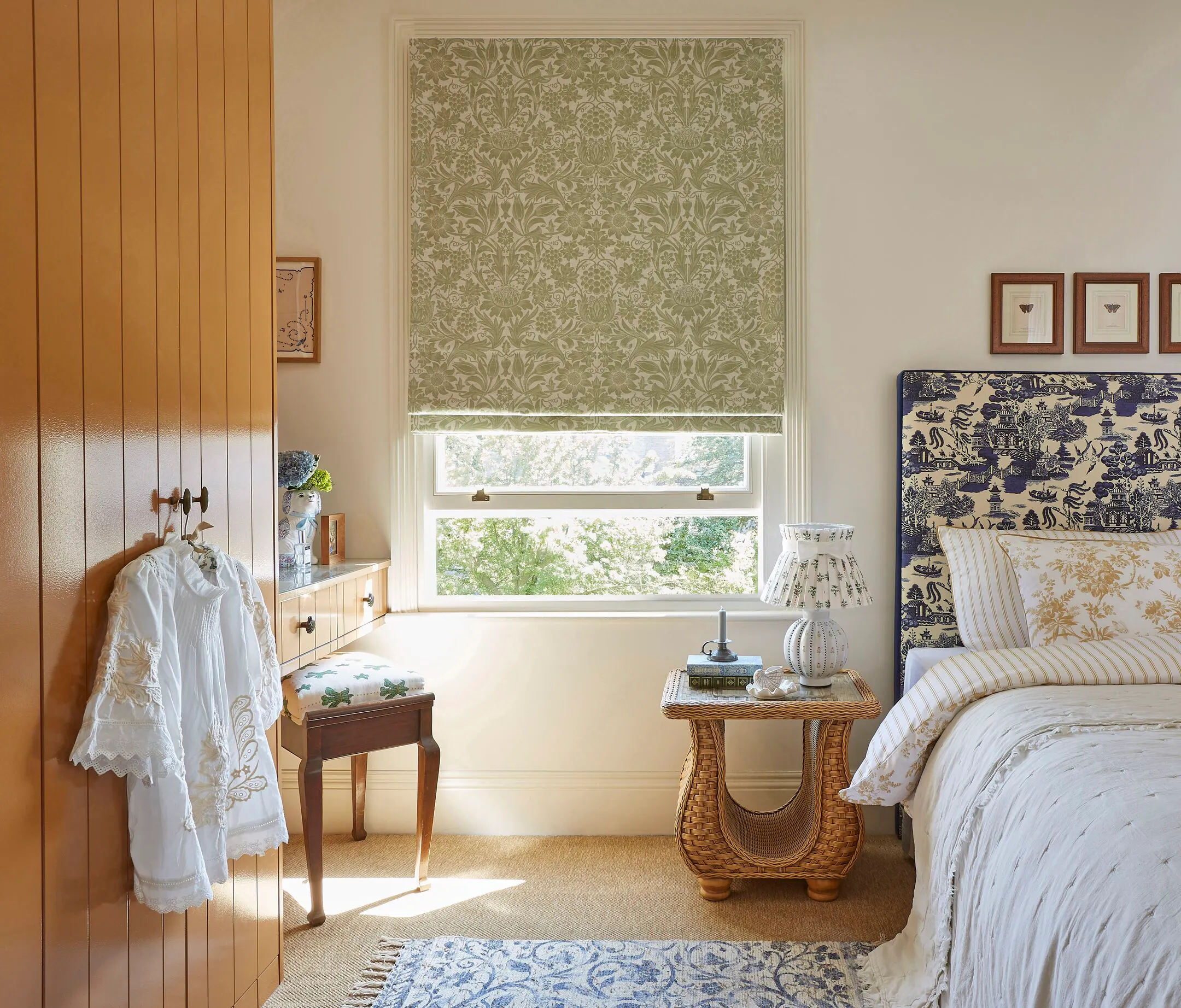 Green patterned Roman blind over a bright bedroom window, with wicker bedside table, upholstered bed and warm timber wardrobe.
