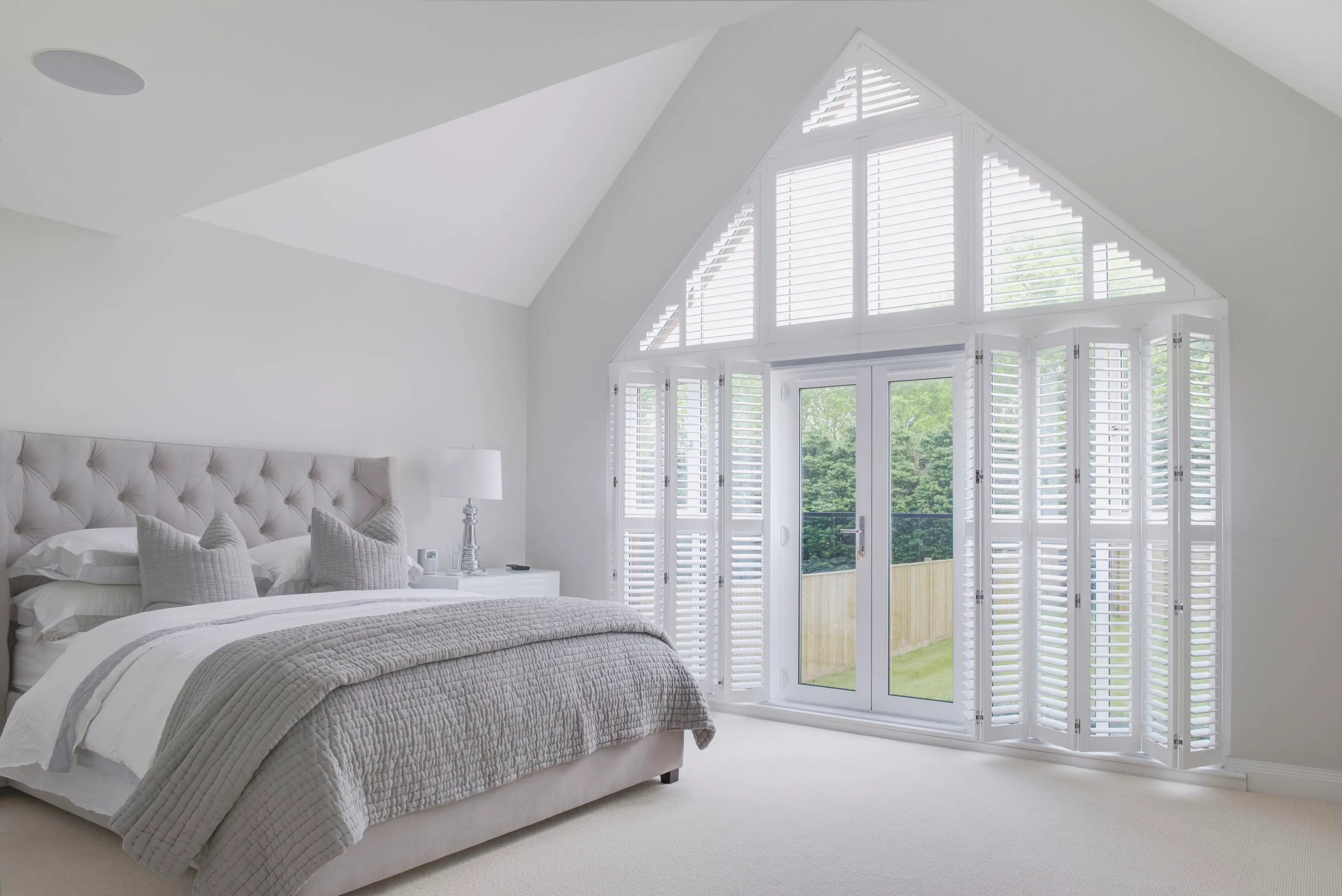 Bright bedroom with a large gable window fitted with white shutters, flooding the calm space with light and garden views.