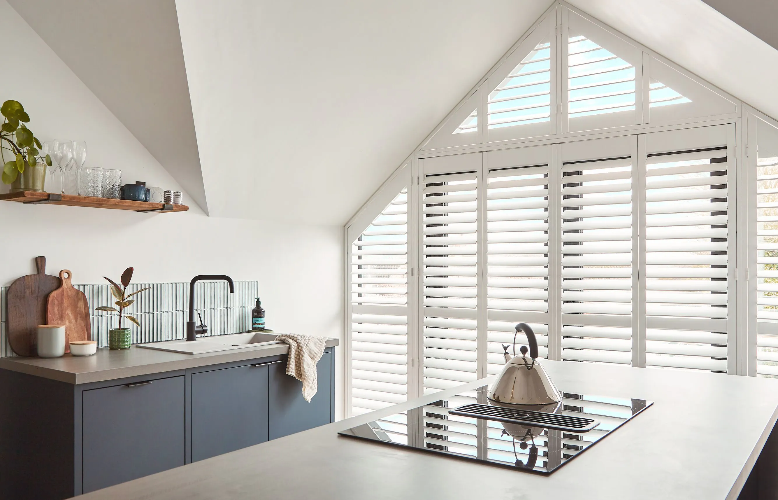 Contemporary kitchen beneath a gable window with white shutters, letting gentle daylight wash across the worktops and hob.