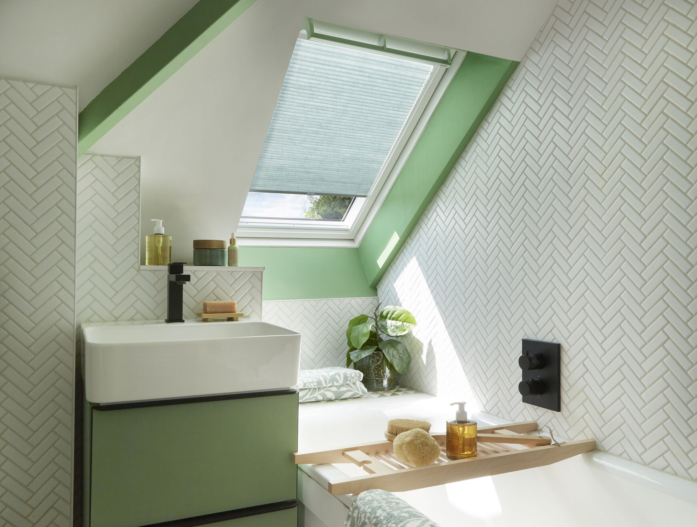 A fresh, green-accented loft bathroom with white herringbone tiles and a green pleated blind fitted to a Velux roof window above the bath.
