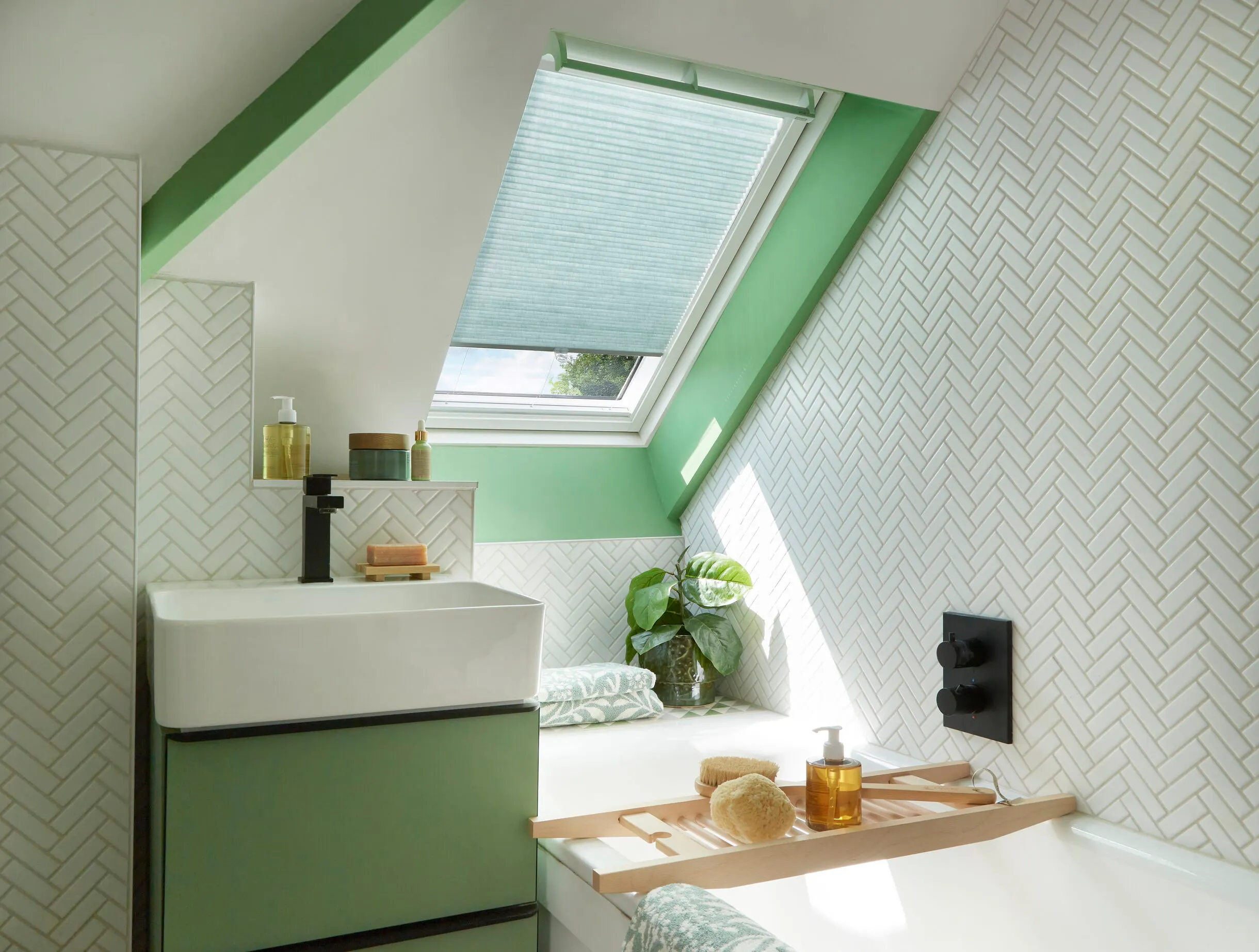 A fresh, green-accented loft bathroom with white herringbone tiles and a green pleated blind fitted to a Velux roof window above the bath.