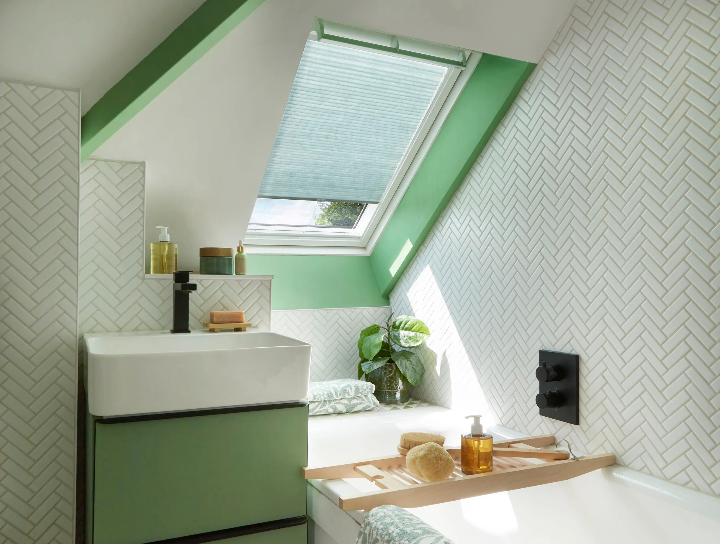 A fresh, green-accented loft bathroom with white herringbone tiles and a green pleated blind fitted to a Velux roof window above the bath.