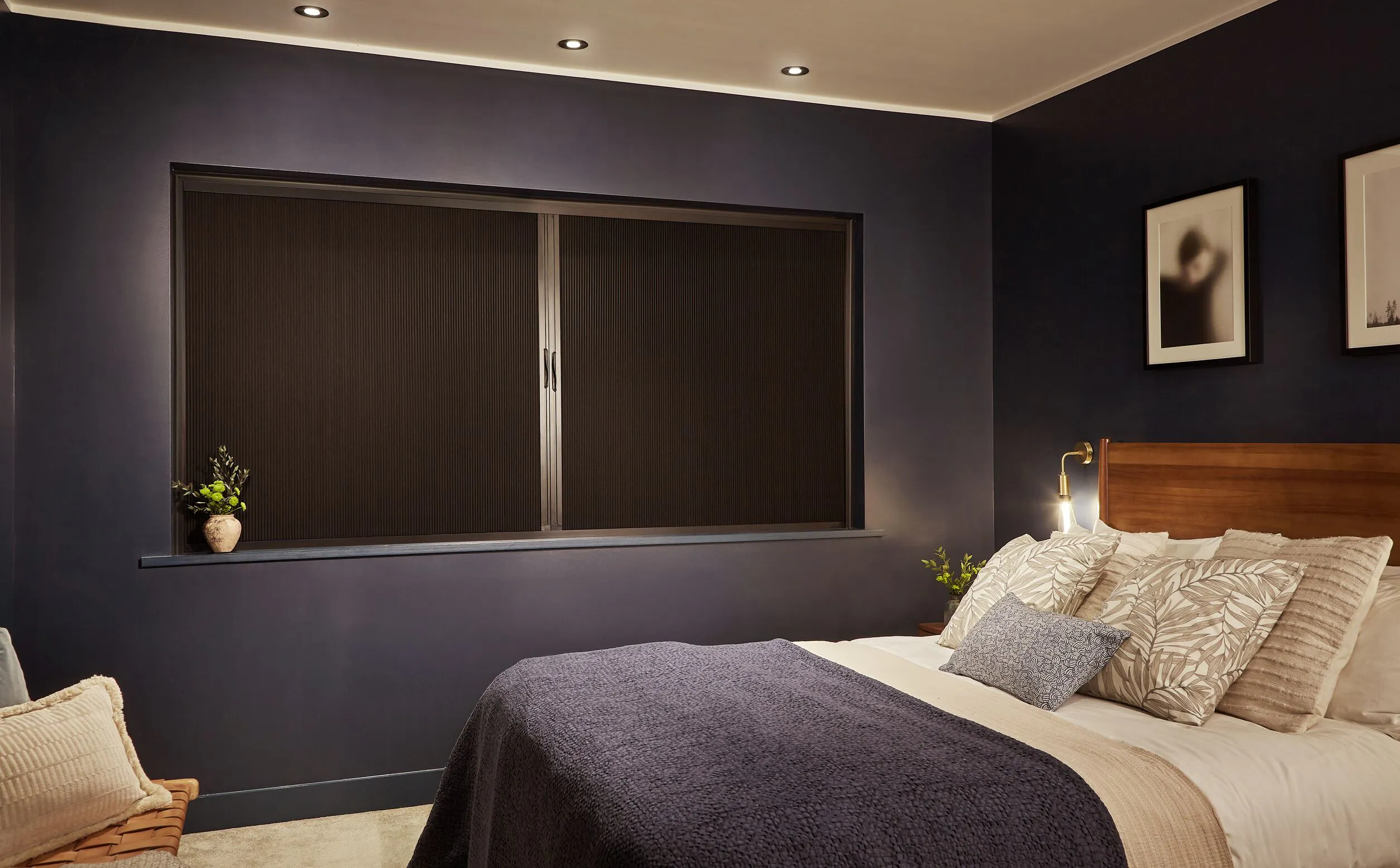 A bedroom with deep navy walls and a large window fitted with a dark Blind Screen, styled with a walnut headboard, layered neutral bedding and warm brass wall lighting.