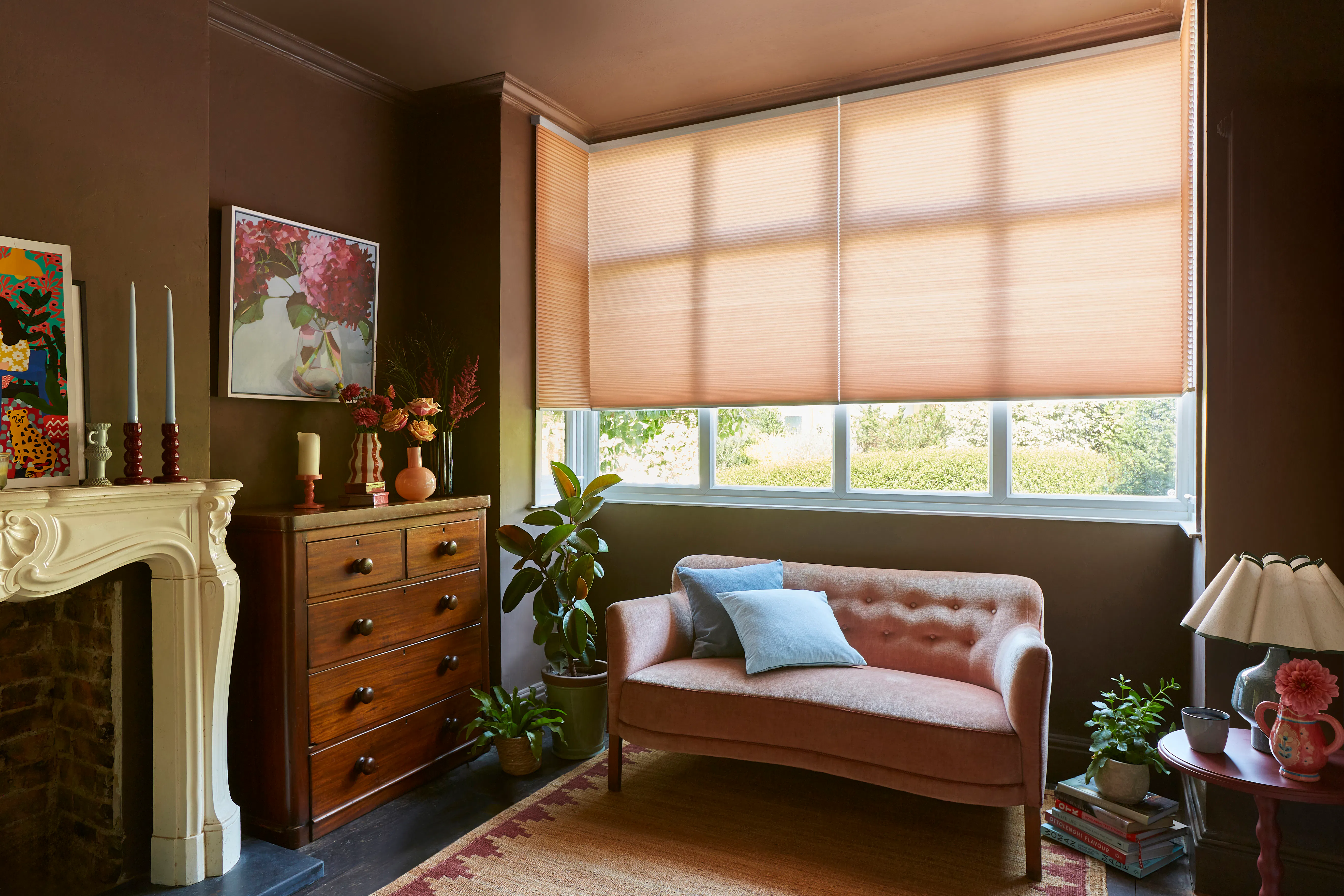  A moody colour-drenched living room with an ornate fireplace and antique chest of drawers. A pink velvet sofa stands beneath a bay window dressed in warm-toned honeycomb blinds. 