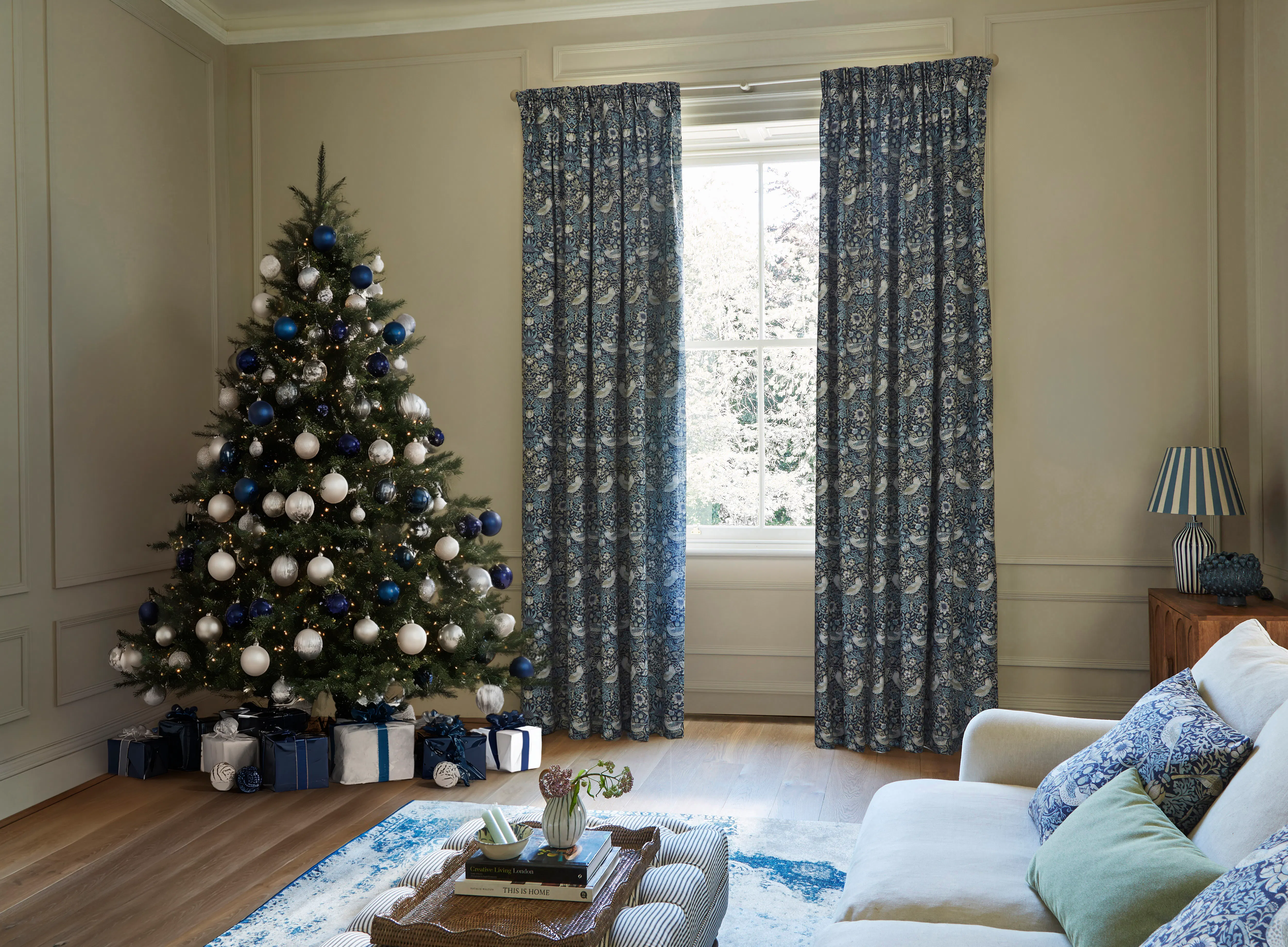 Christmas tree with blue and white decorations beside a large window framed by patterned blue curtains, with wrapped gifts beneath.