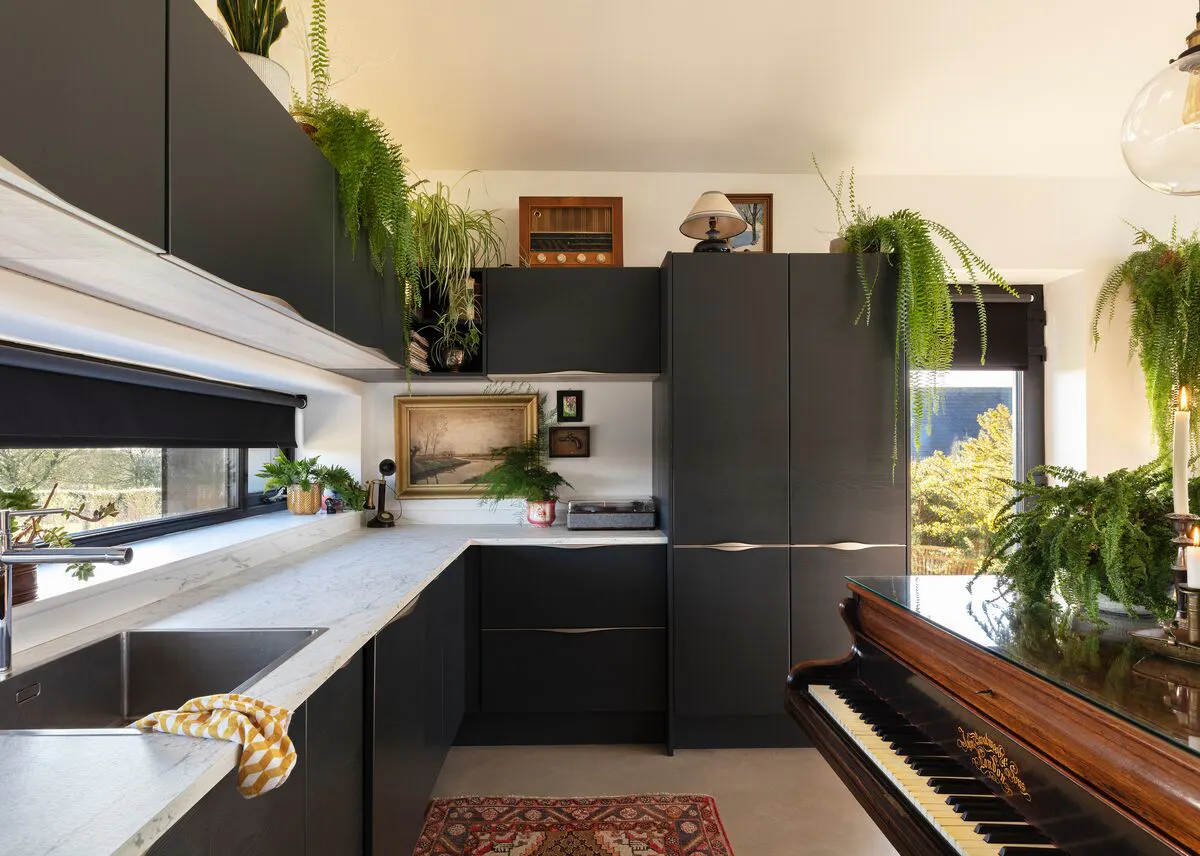 A moody black kitchen with marble countertops, a grand piano and trailing houseplants.