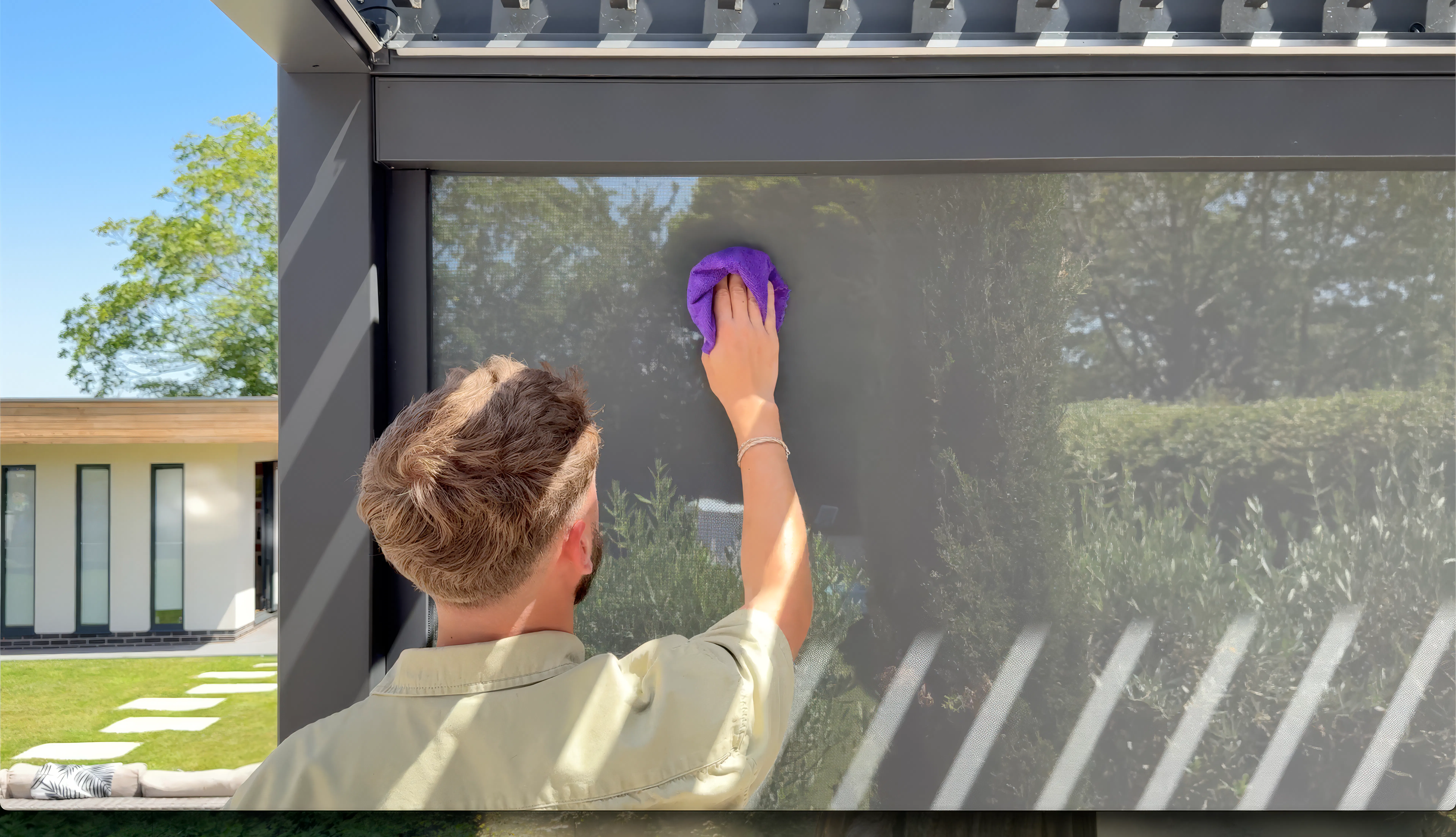 Person cleaning a pergola side screen with a purple cloth beneath a modern outdoor structure in a sunny garden.
