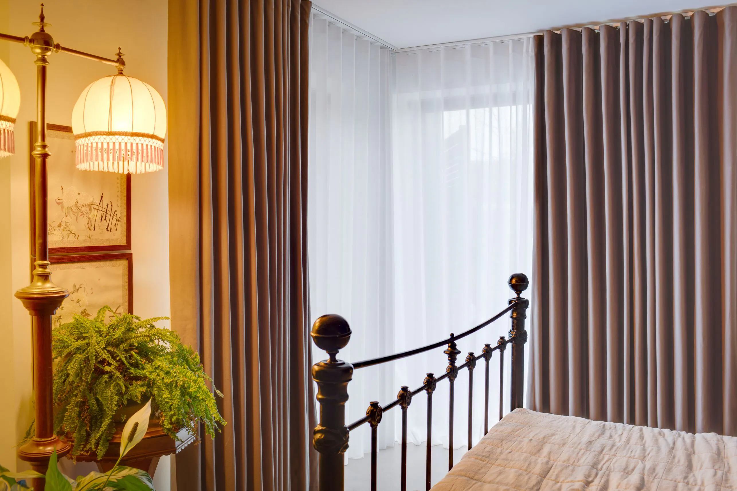 Bedroom with tall window dressed in sheer curtains and grey drapes, softly diffusing daylight beside an iron bedframe and leafy plant.