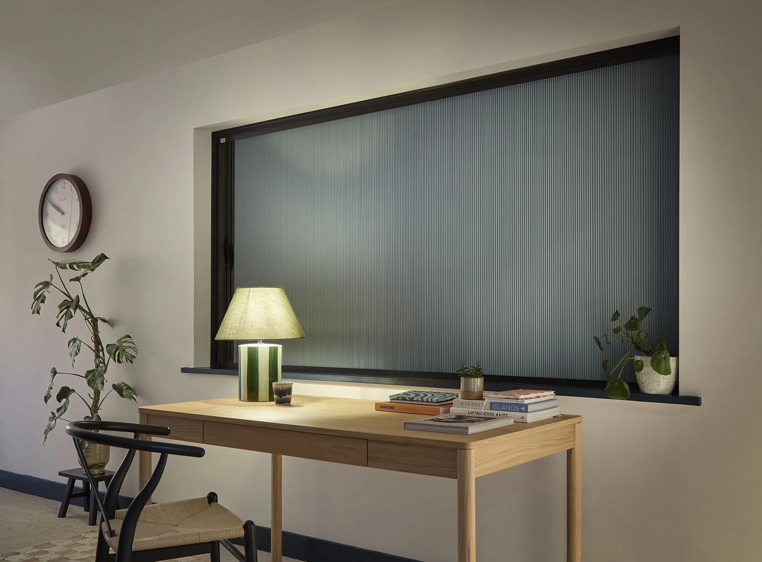 Pale Scandi-style desk in front of a large black-framed window, entirely screened by a blue-grey pleated blind. Styled with a green lamp and houseplants.