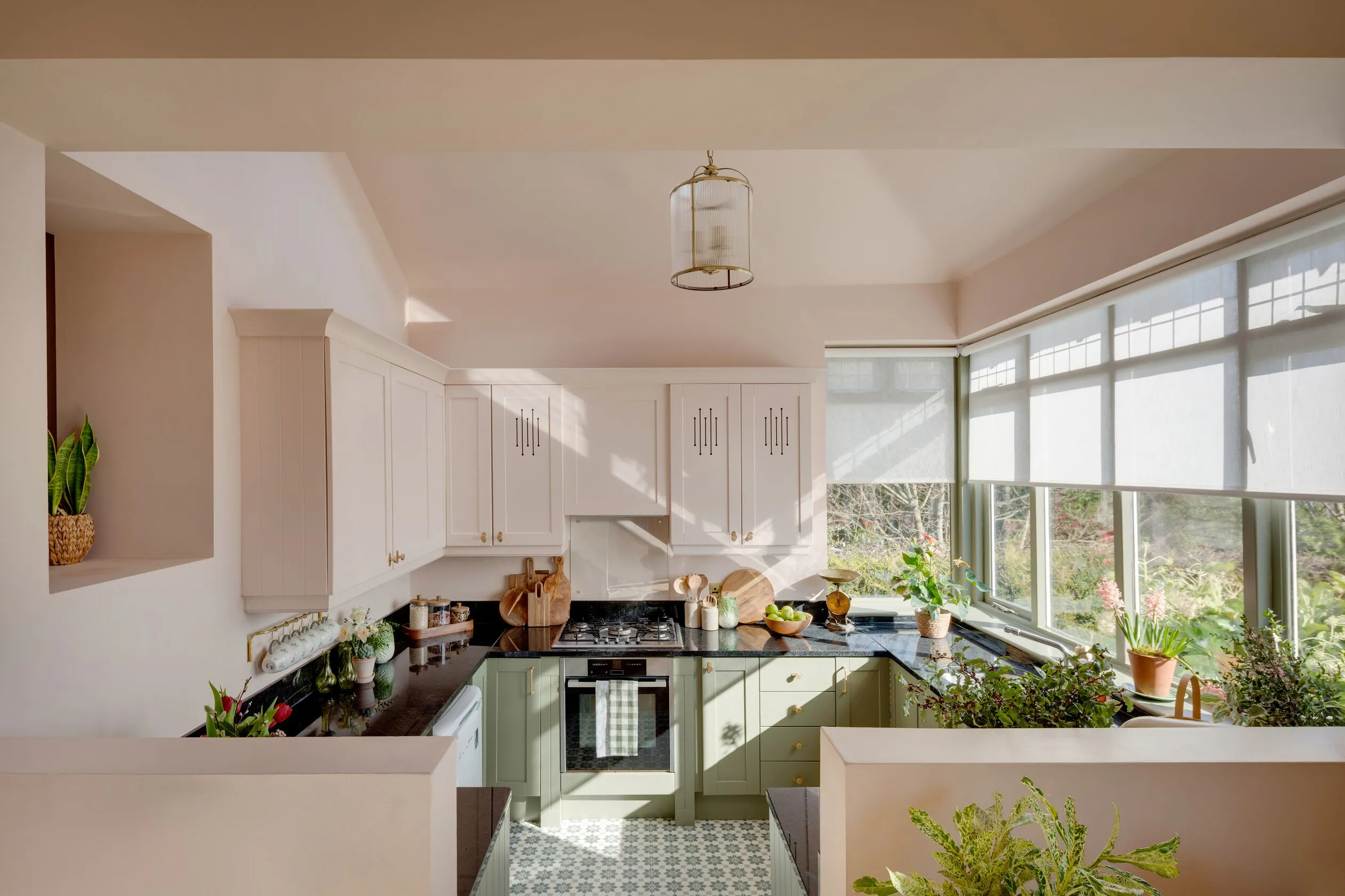Light-filled kitchen with wide windows fitted with Roller blinds, overlooking greenery and filling the space with natural light above sage cabinets.
