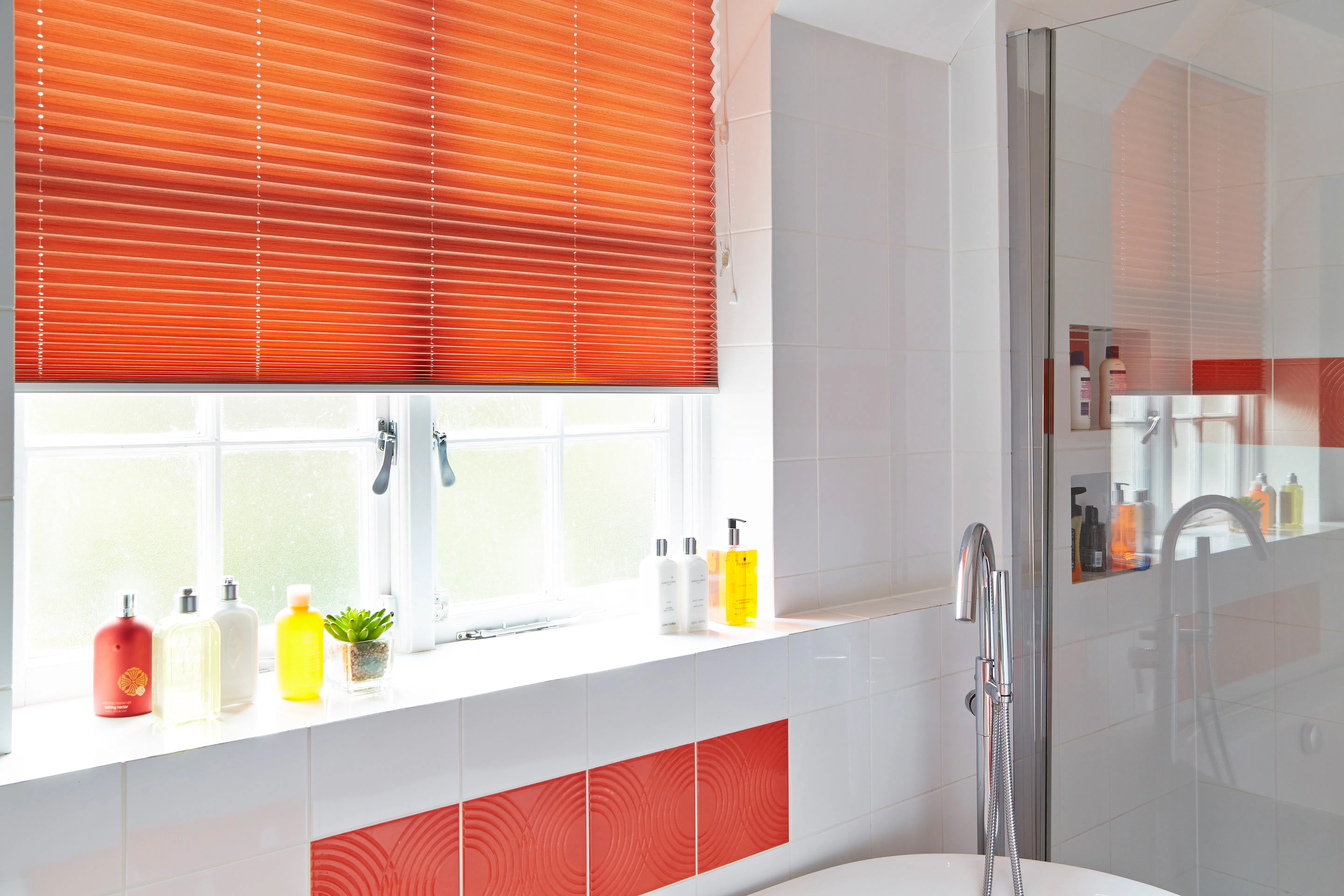 Pleated blind in a bold orange shade covering a bathroom window, adding colour to the white tiled space with bath and shower.