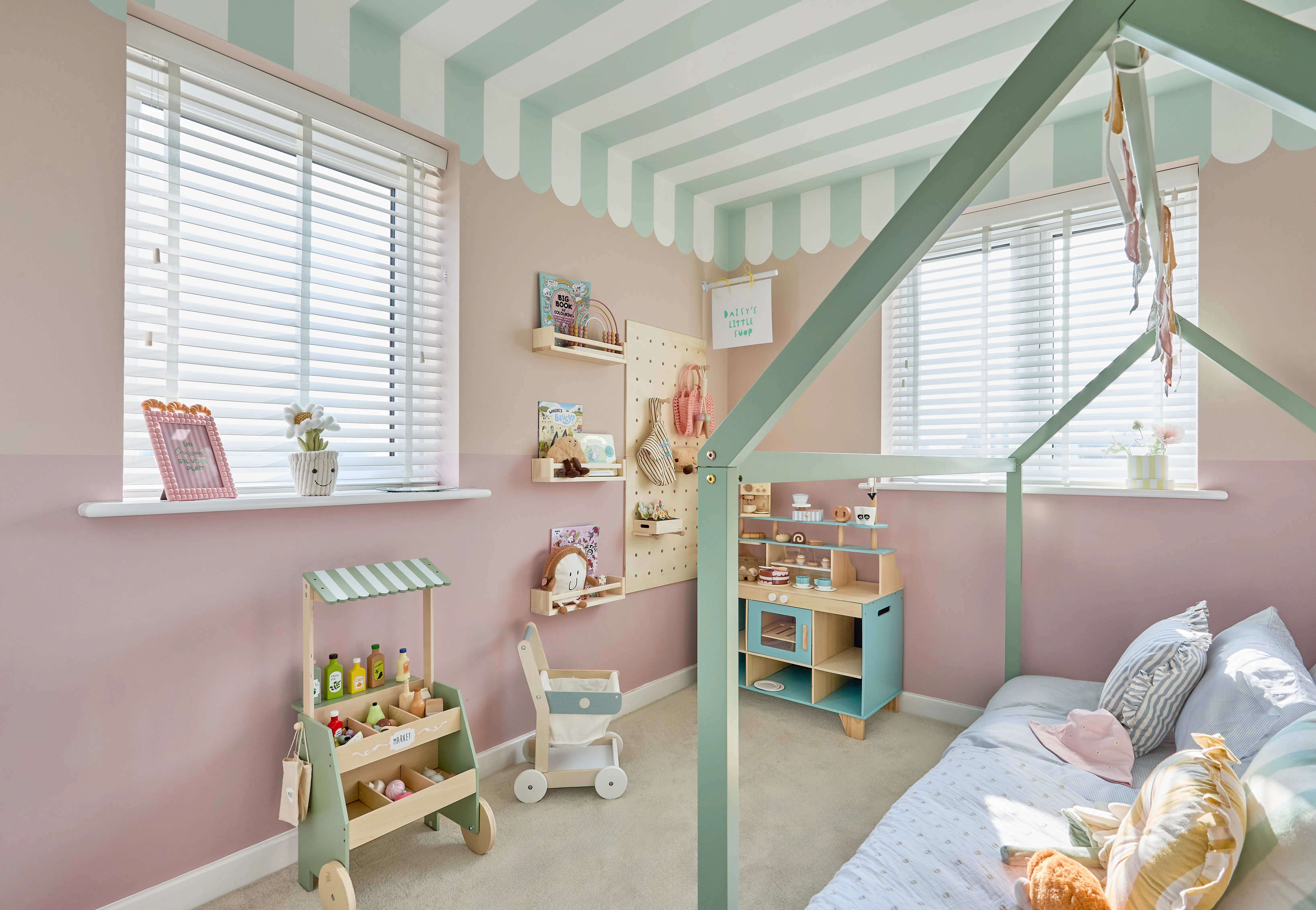 A pastel pink and sage green playroom with a striped canopy ceiling, wooden play shop and Wooden blinds.