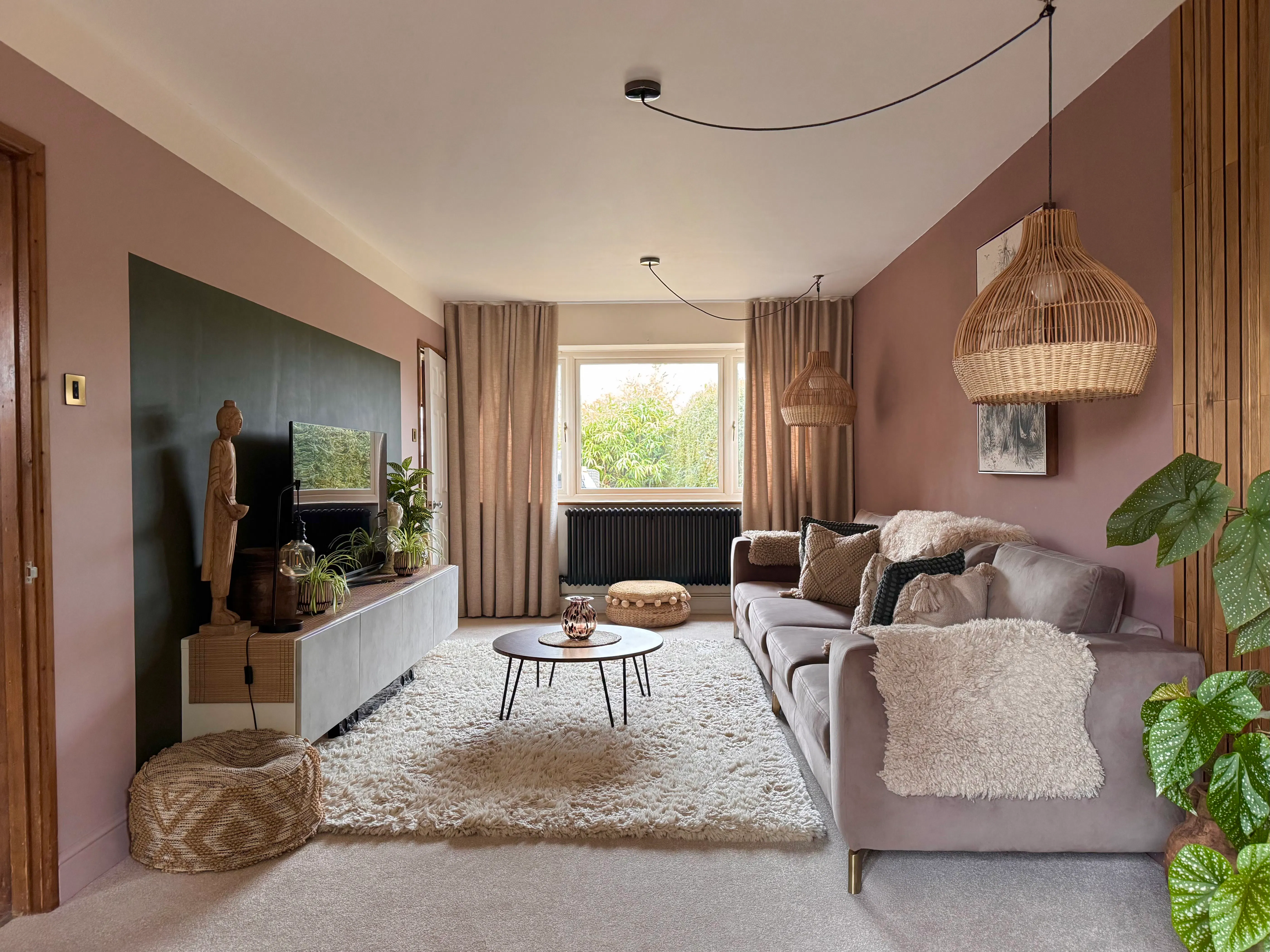 Long living room with central window dressed in soft curtains, grey sofa along the wall, shaggy rug, round coffee table and warm woven pendant lights.