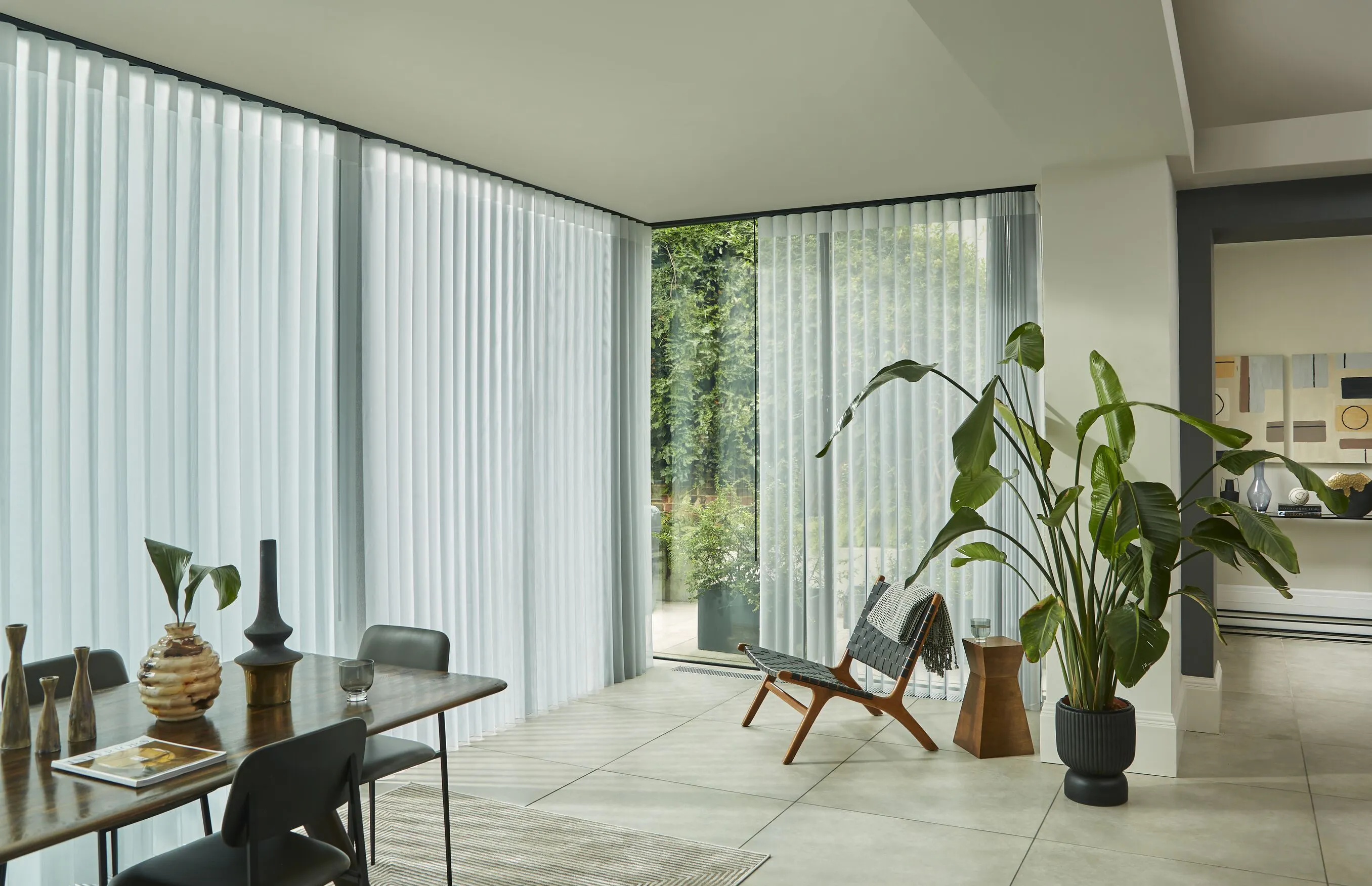 Corner glazing fitted with white Vertical blinds, filtering daylight into a modern dining area with large indoor plants and garden views.