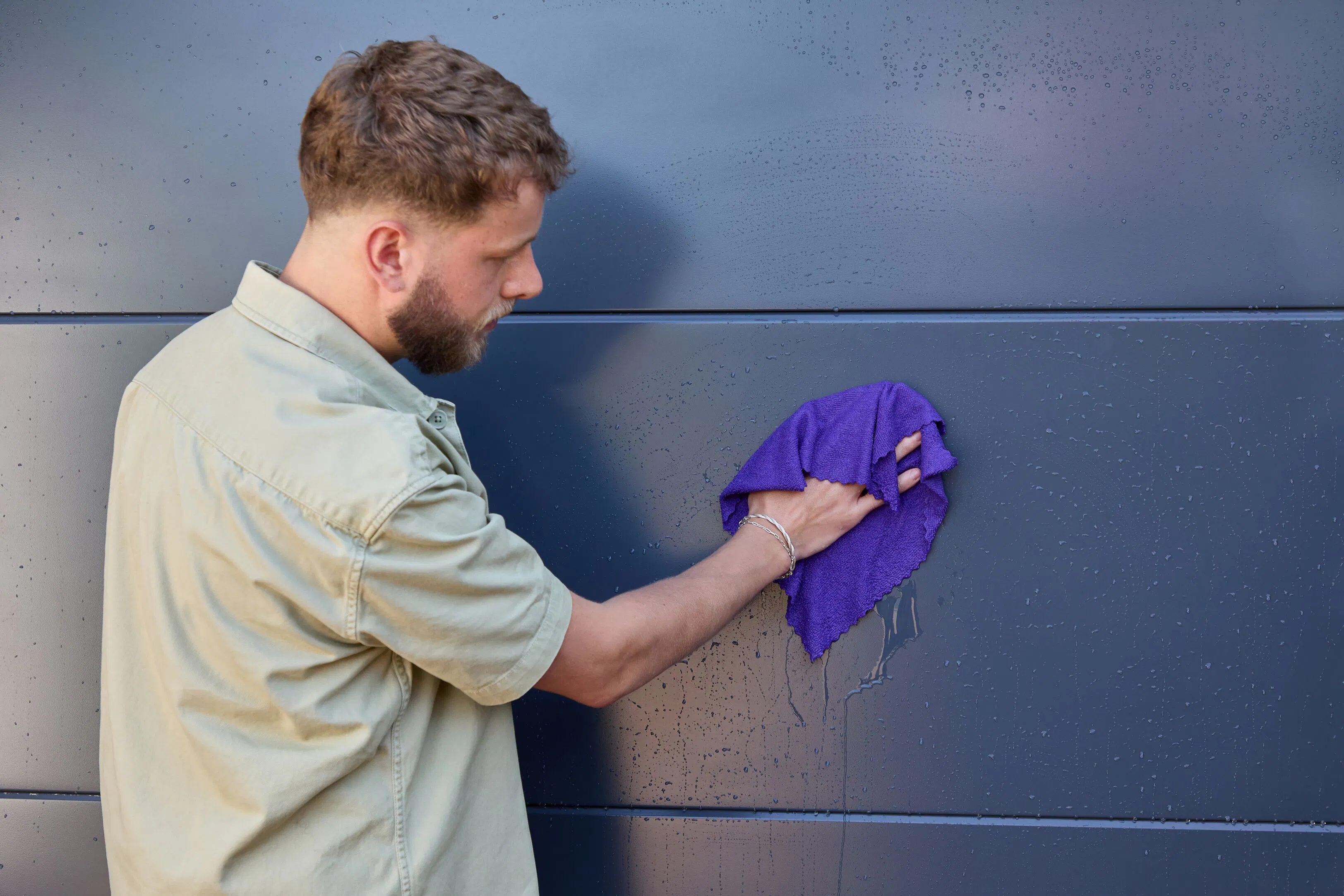 Person wiping down an anthracite grey garage door with a purple cloth, removing water droplets from the smooth panelled surface.