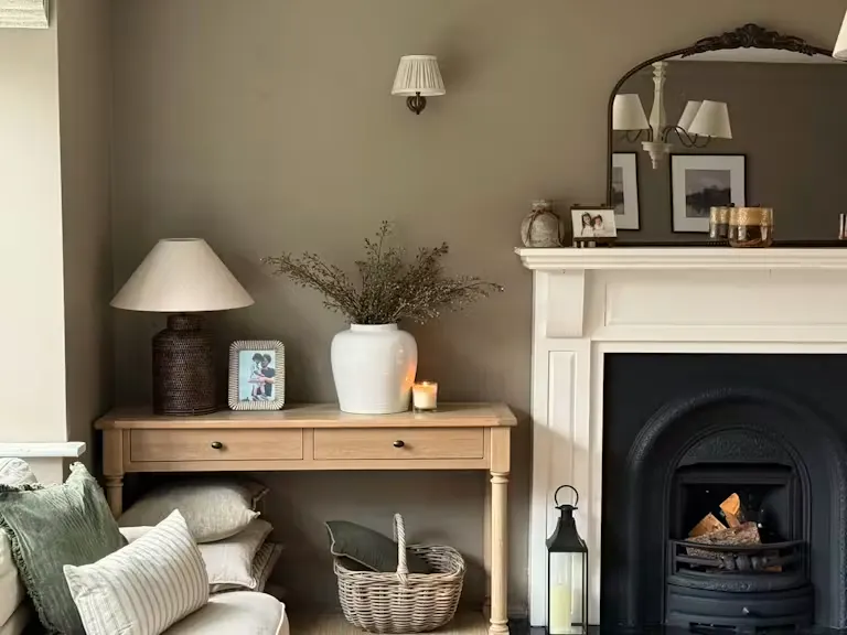 Cosy living room corner with a wooden console table, ceramic vase, lamp and candle beside a classic fireplace with mirror and framed photos.