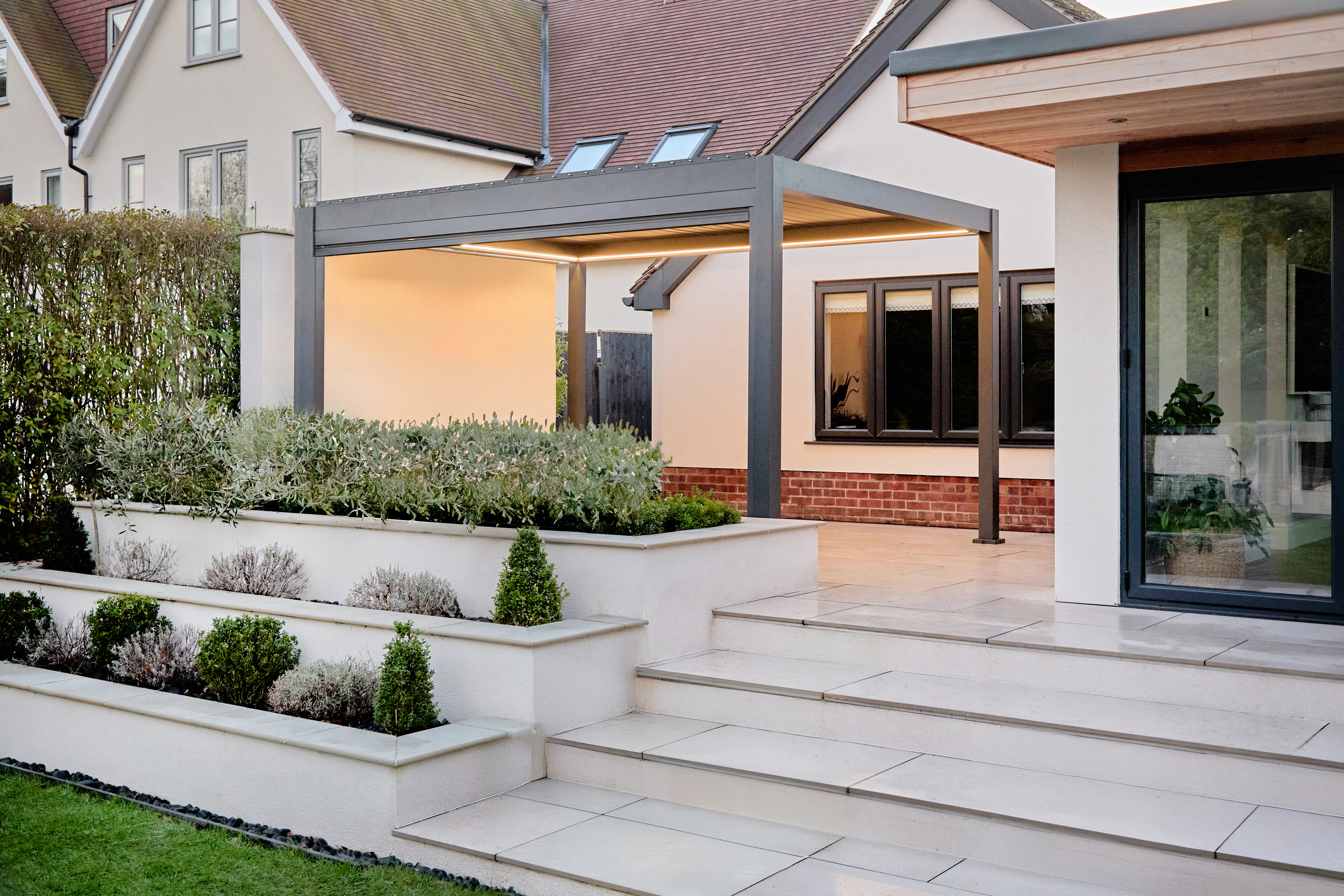 Evening view of a dark grey louvred pergola with integrated lighting above stone steps and raised planted borders.