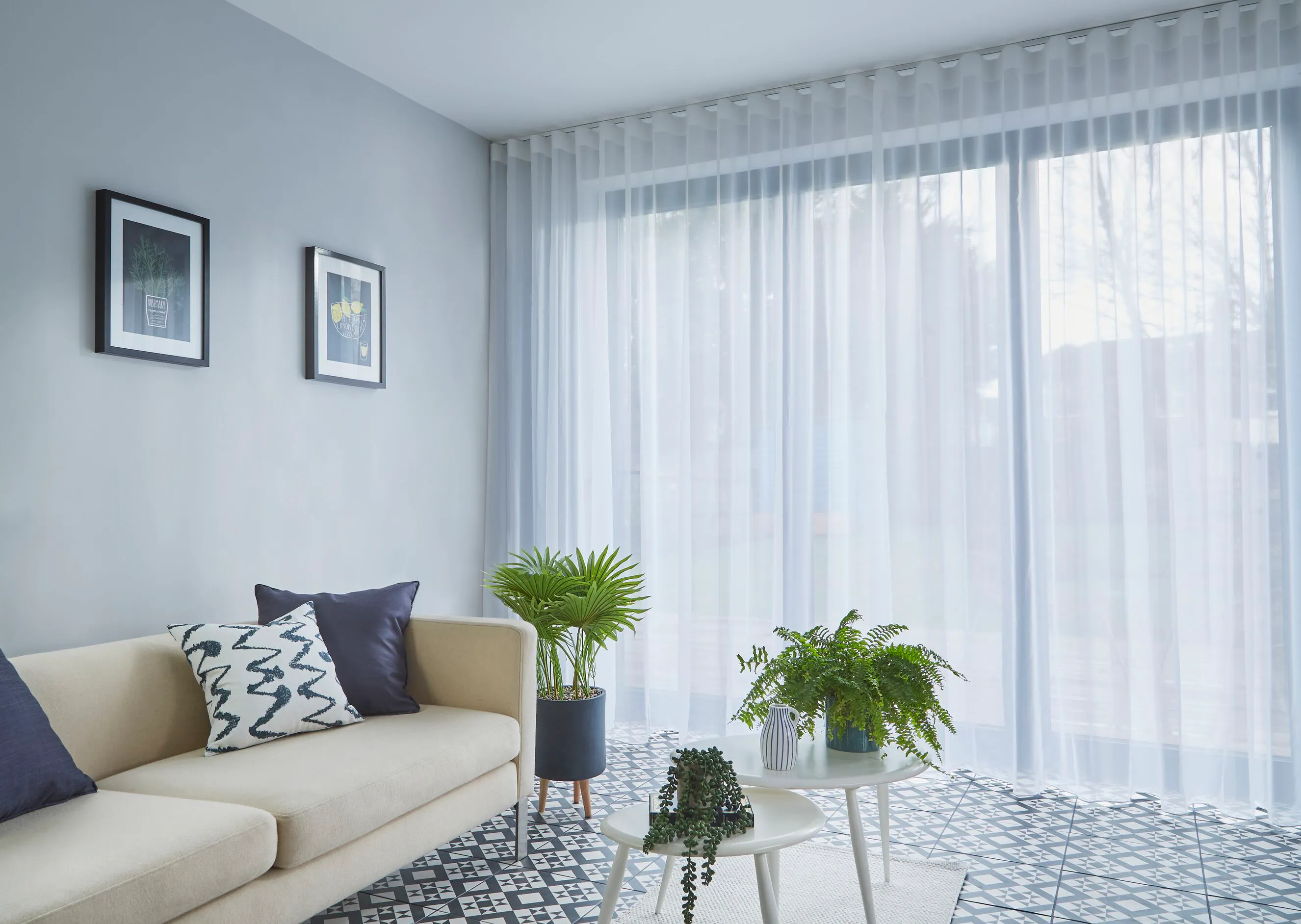 A pale blue-grey room with a cream sofa, potted plants, patterned floor tiles and soft light filtering through floor-to-ceiling sheer curtains.