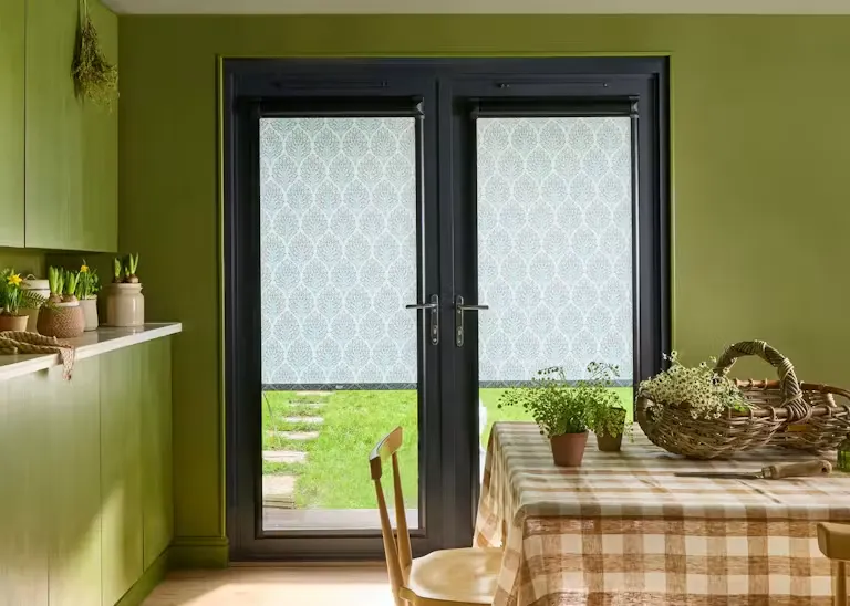 Dark-framed patio doors fitted with patterned Perfect Fit Roller blinds, letting soft light into a green kitchen-diner overlooking the garden.