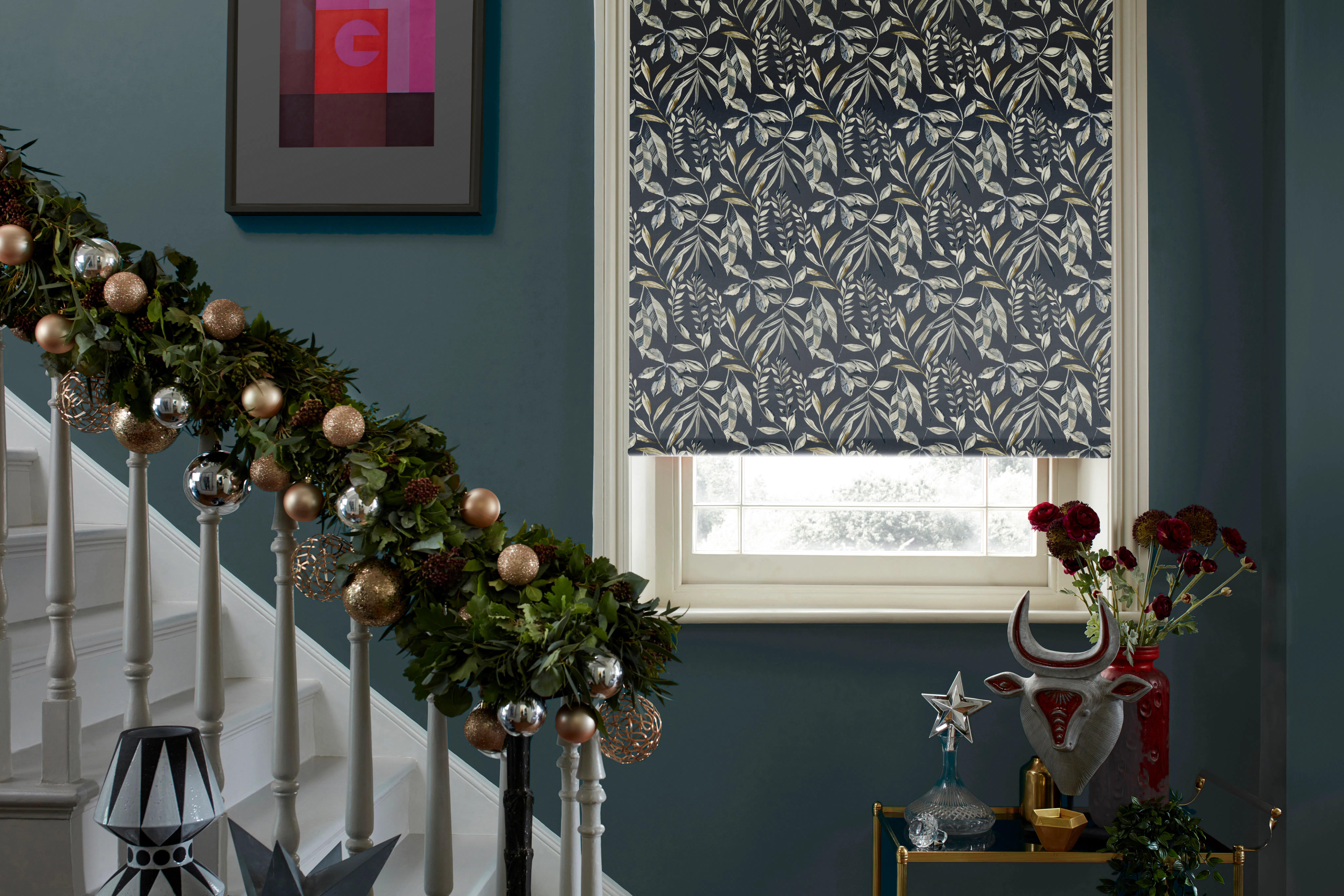 A blue-grey stairway with a sash window dressed with a navy leaf-print Roman blind. The banister is wrapped in a gold and silver bauble garland, and a small gold-toned console table is styled with deep red flowers. 